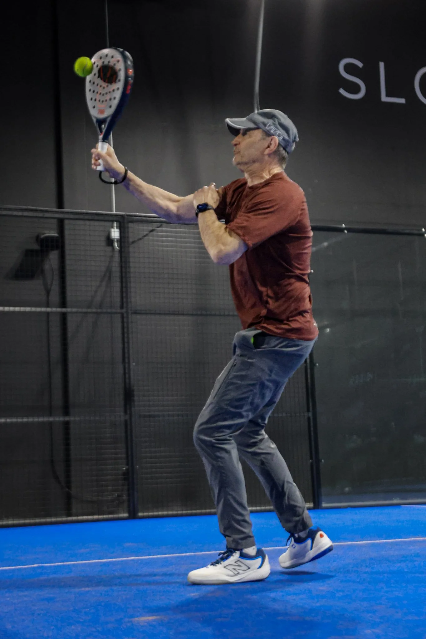 A man wearing a gray cap, a rust-colored t-shirt, gray pants, and white sneakers playing indoor padel, preparing to hit a ball with a padel racquet on a blue padel court with black walls.