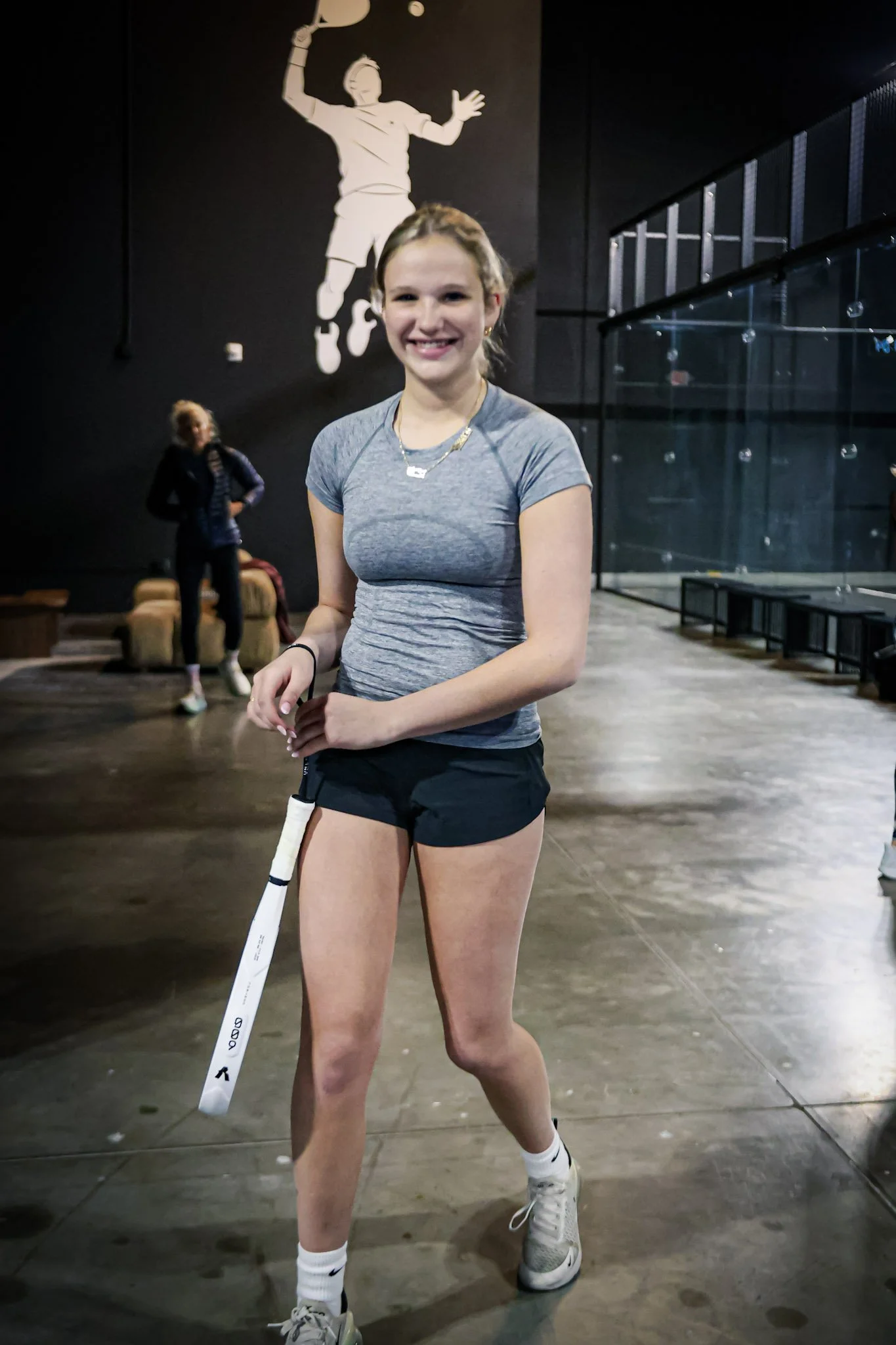 A young woman smiling in a well-managed padel club holding a white stick or pole, with a black wall featuring a large white image of a padel player in the background.