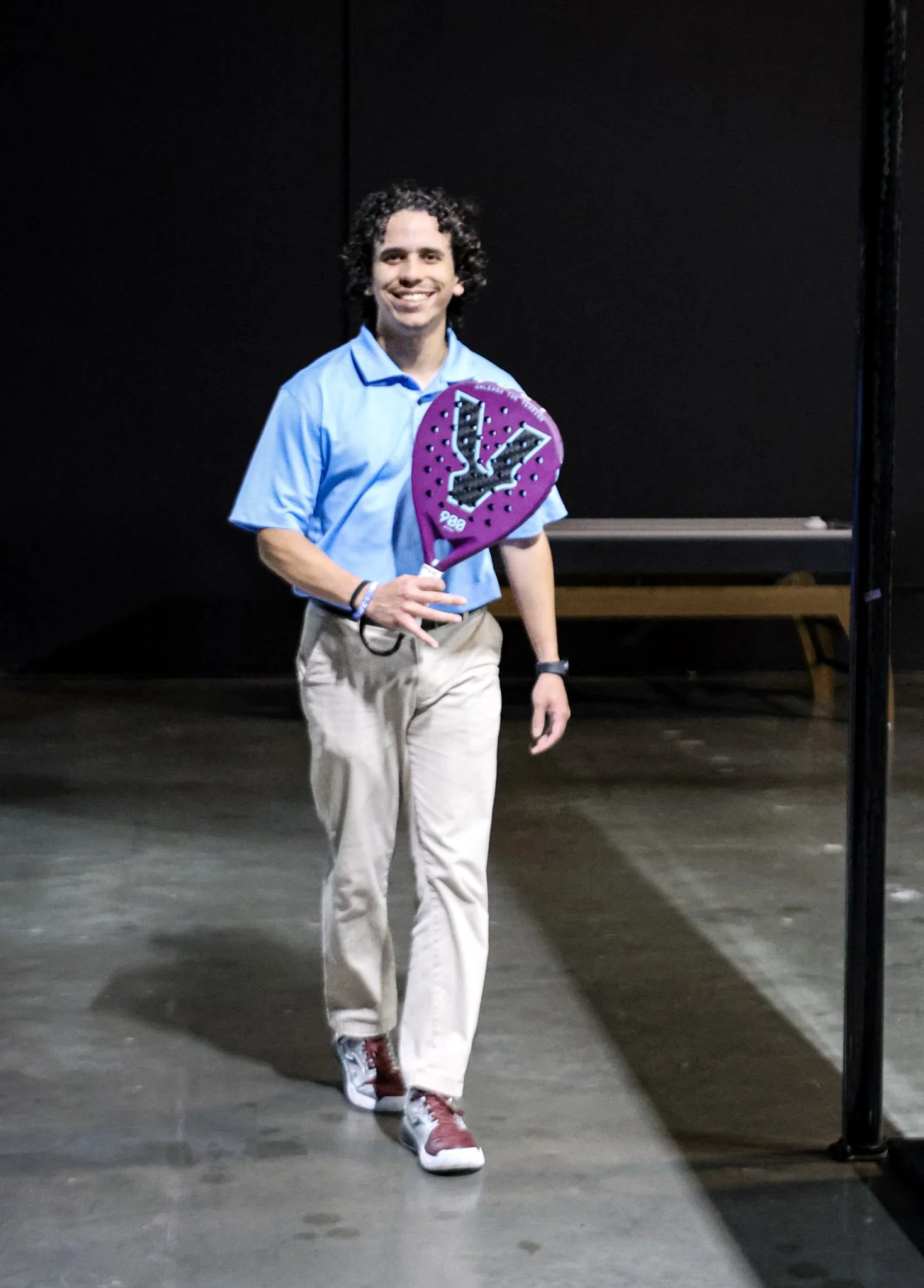 A man with curly dark hair smiling, wearing a light blue polo shirt, beige pants, and red sneakers, holding a purple padel racquet, standing indoors against a black background.