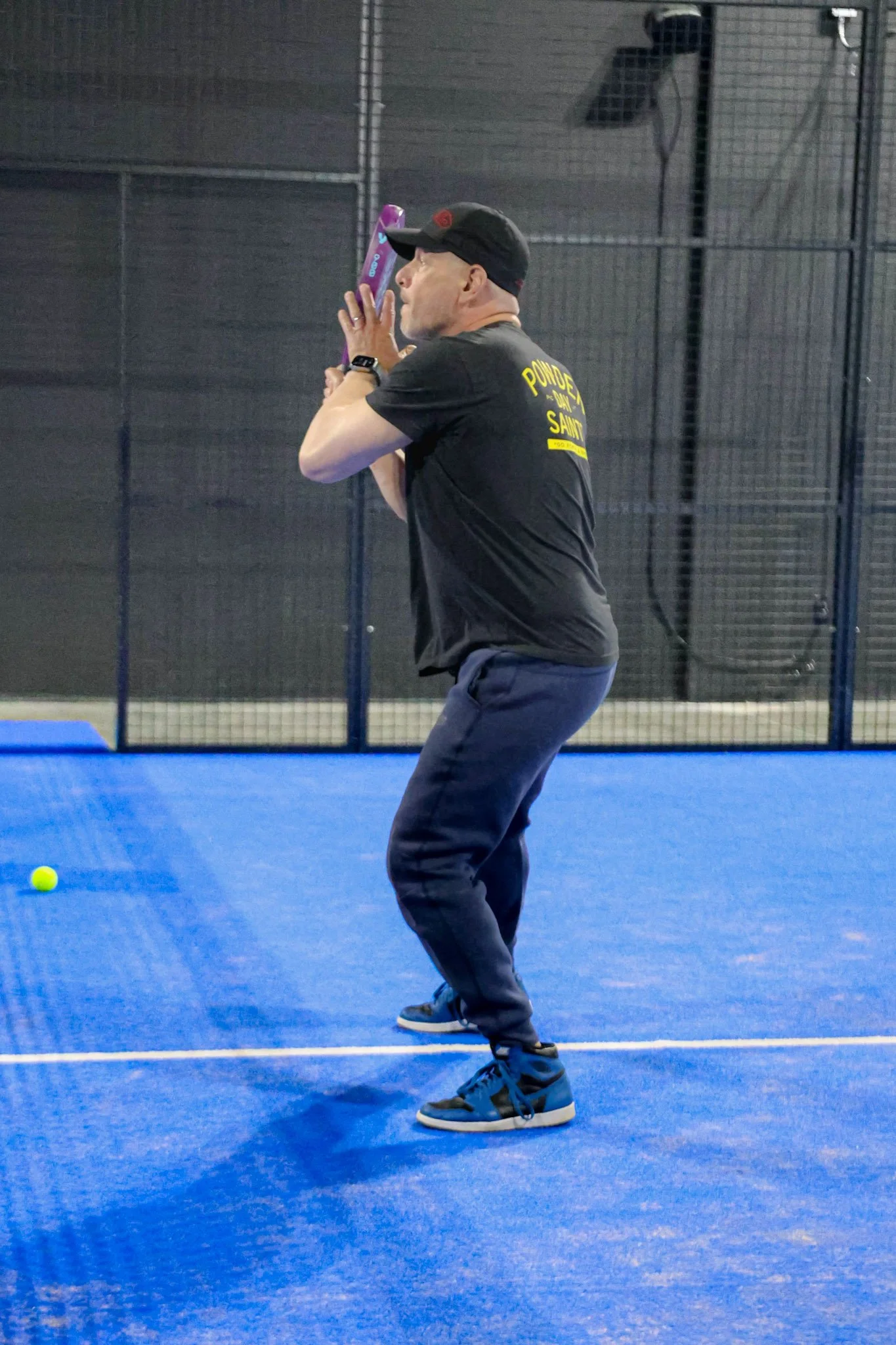 A man playing paddle tennis on a blue surface, wearing a black cap, black T-shirt with yellow text, gray pants, and athletic shoes, holding a padel racquet and preparing to hit a yellow padel ball.