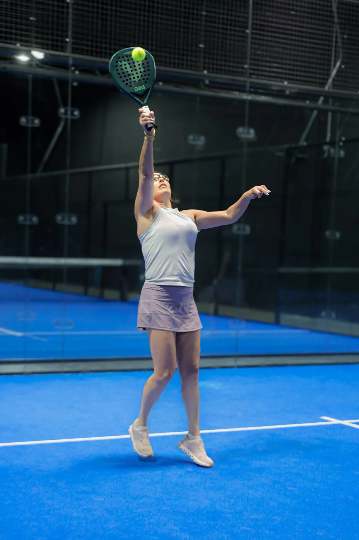 A woman playing padel on an indoor padel court, preparing to hit a bright yellow padel ball with a padel racquet. She is wearing sunglasses, a sleeveless top, a skirt, and athletic shoes.
