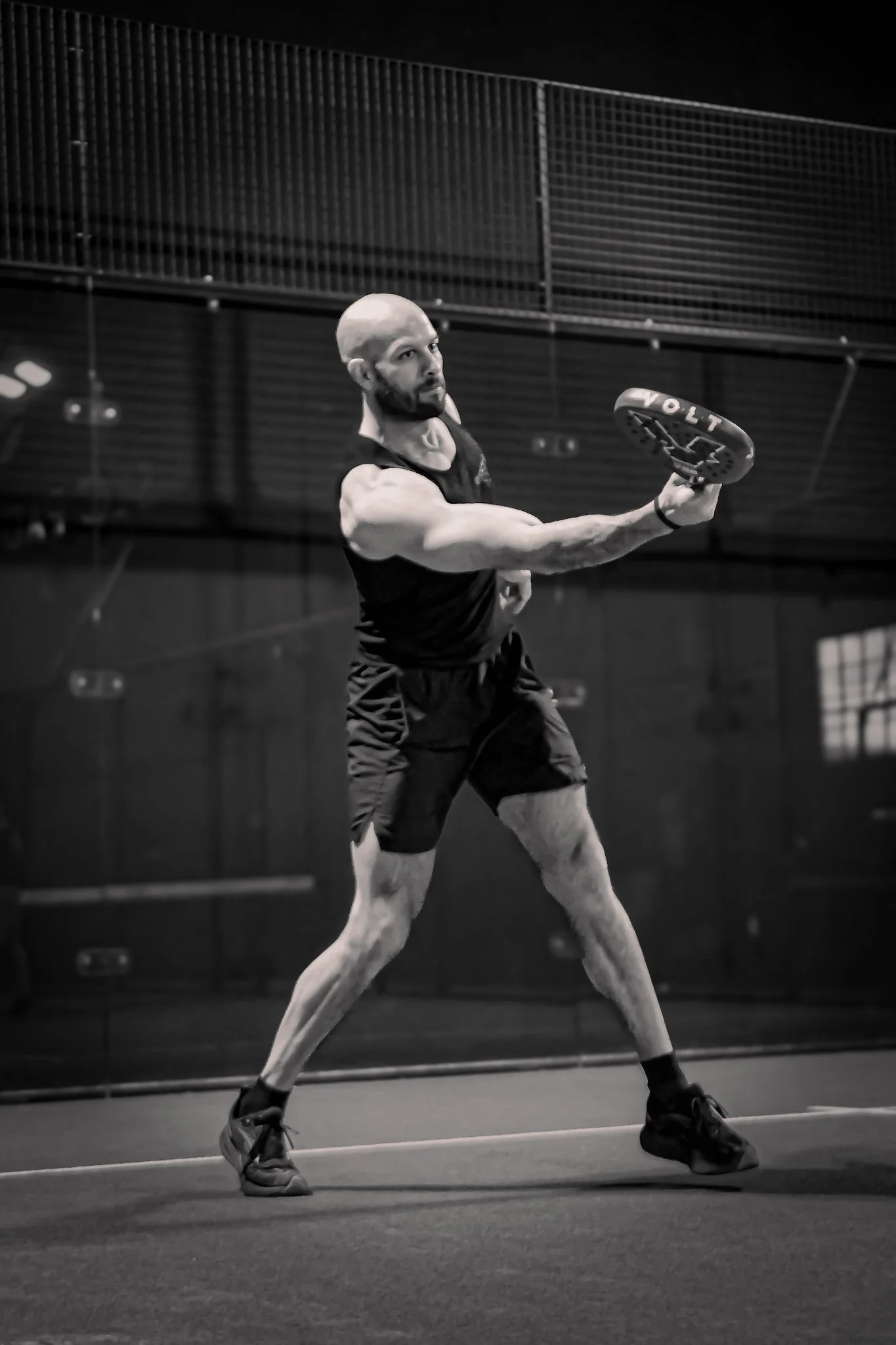 A muscular man wearing a sleeveless sports shirt and shorts, is playing padel indoors. He is holding a padel racquet and appears to be about to hit a padel ball. The background features a high ceiling with netting or fencing.