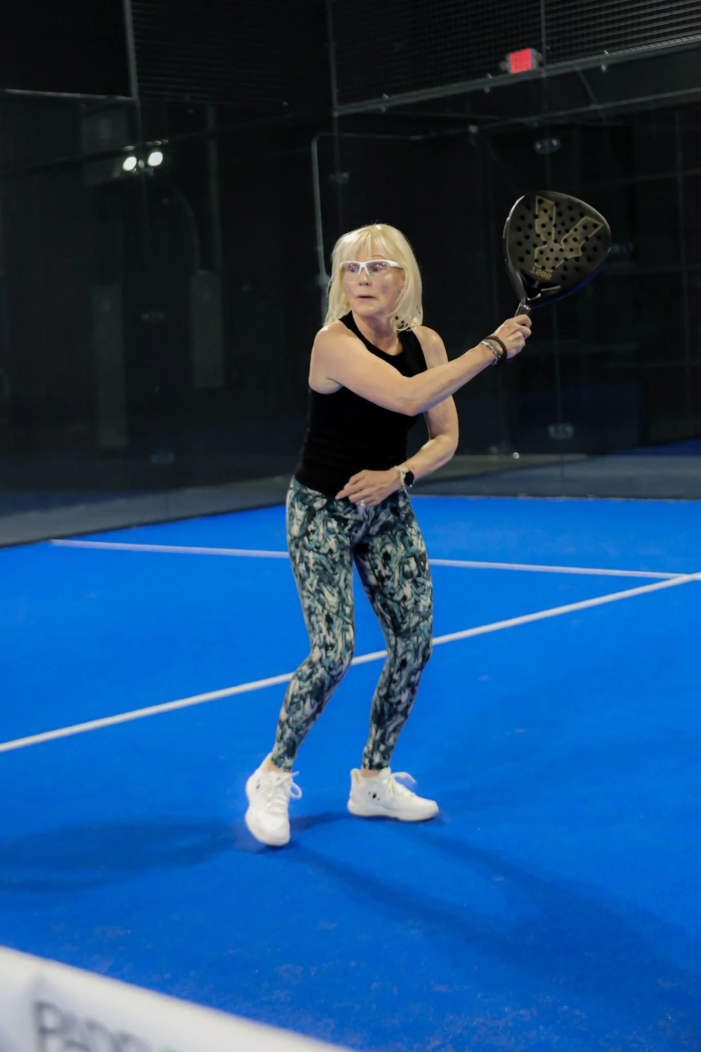 An older woman with blonde hair, glasses, wearing a black sleeveless top, patterned leggings, and white sneakers, is playing padel indoors, holding a padel racket in a ready position.