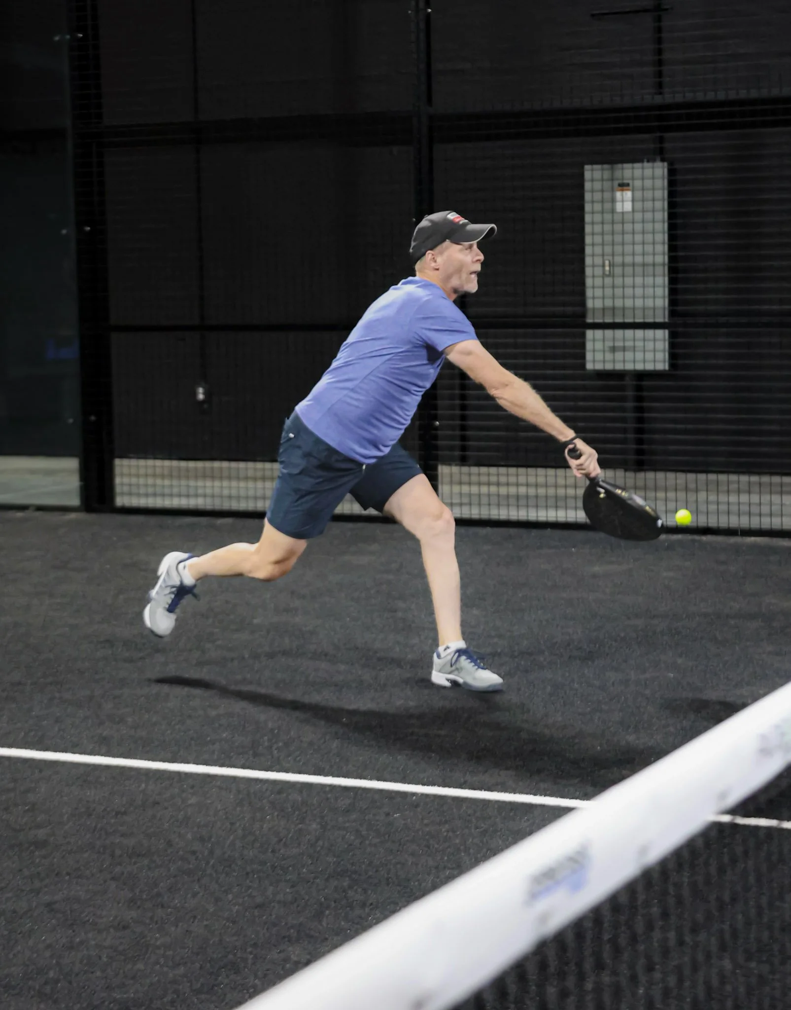 A man wearing a black cap, blue shirt, and black shorts playing padel indoors on a black padel court, preparing to hit a padel ball with a padel racquet, with a black background and a white line on the court.