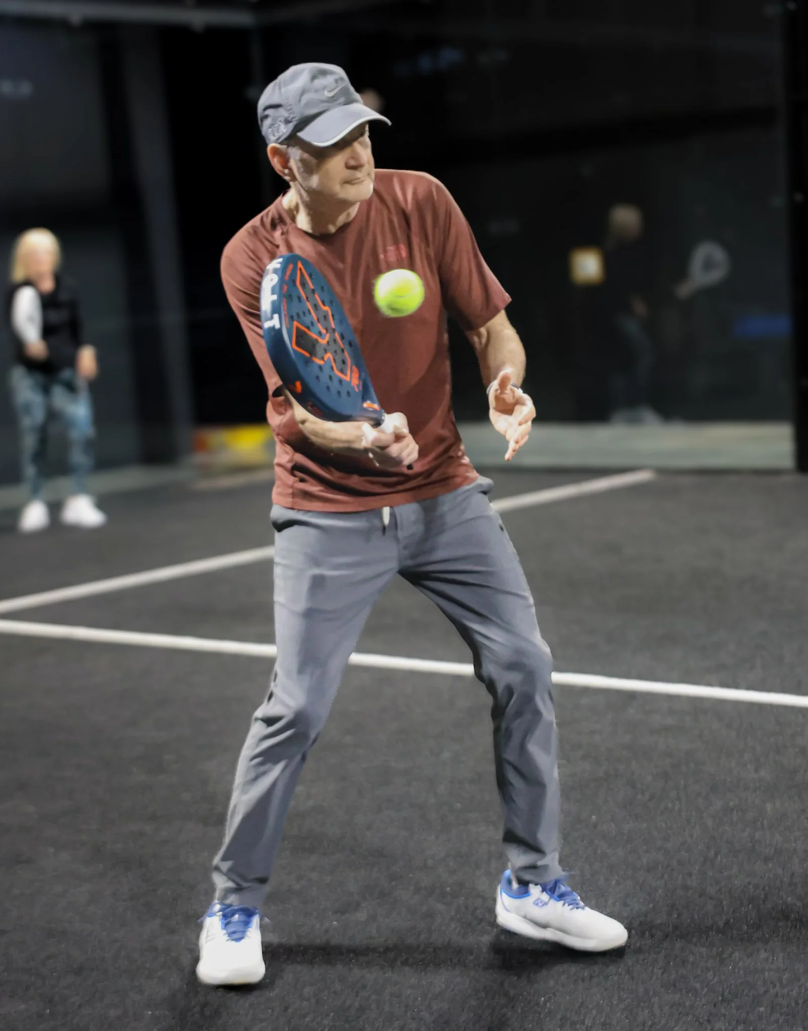 A man wearing a gray cap, burgundy T-shirt, gray pants, and white athletic shoes with blue accents playing padel, about to hit a yellow padel ball on an indoor court with black flooring.