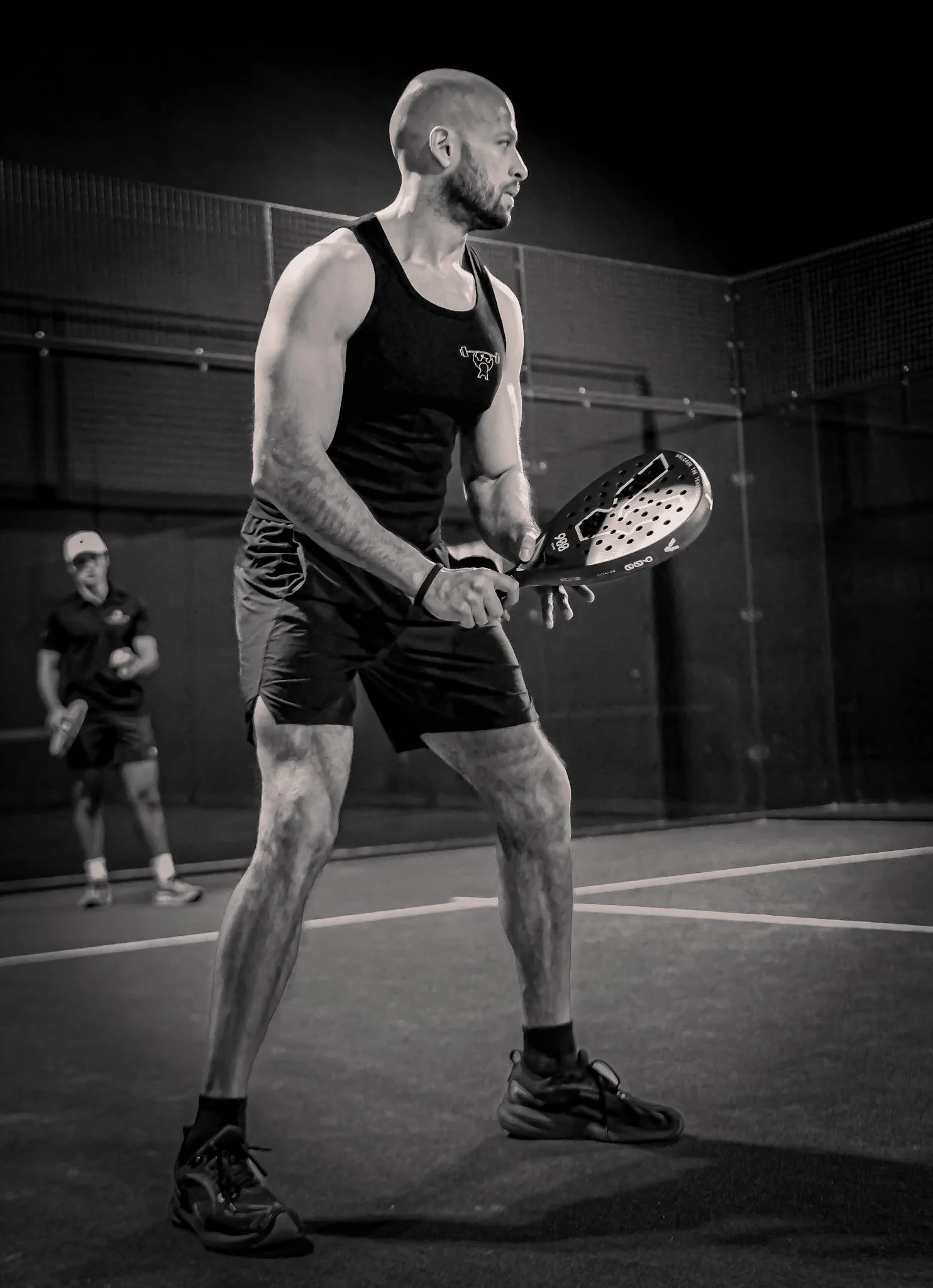 A man playing padel on an indoor padel court, holding a padel racquet, with a woman watching in the background.