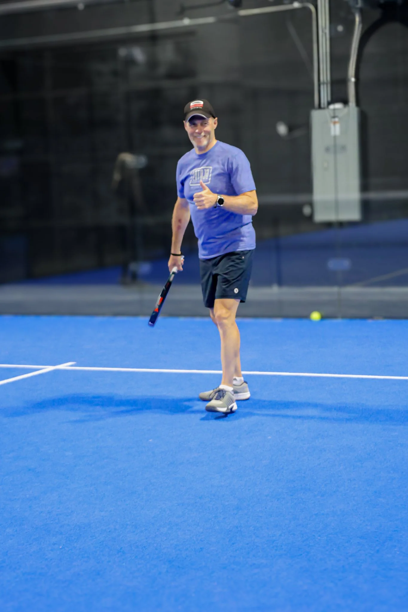 A man smiling and giving a thumbs-up on a blue indoor padel court, holding a padel racquet, with a padel ball on the ground nearby, wearing a blue T-shirt, black shorts, a cap, and an athletic watch.