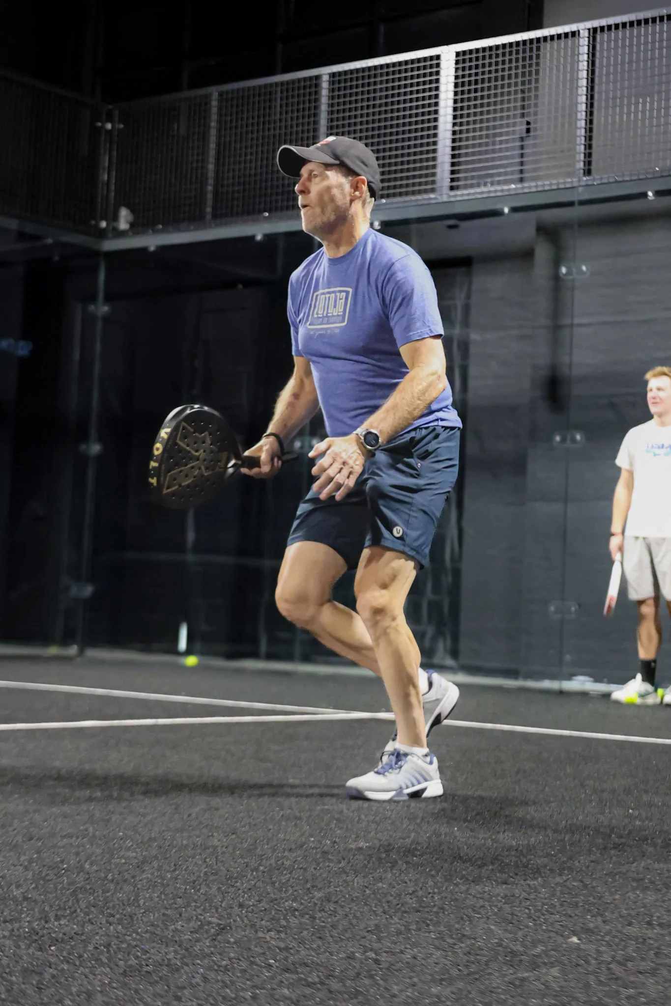 A middle-aged man in athletic clothing holding a padel racquet, running on an indoor padel court. He is wearing a black cap, blue T-shirt, shorts, and sneakers. In the background, there is a young man in white standing near the padel court.
