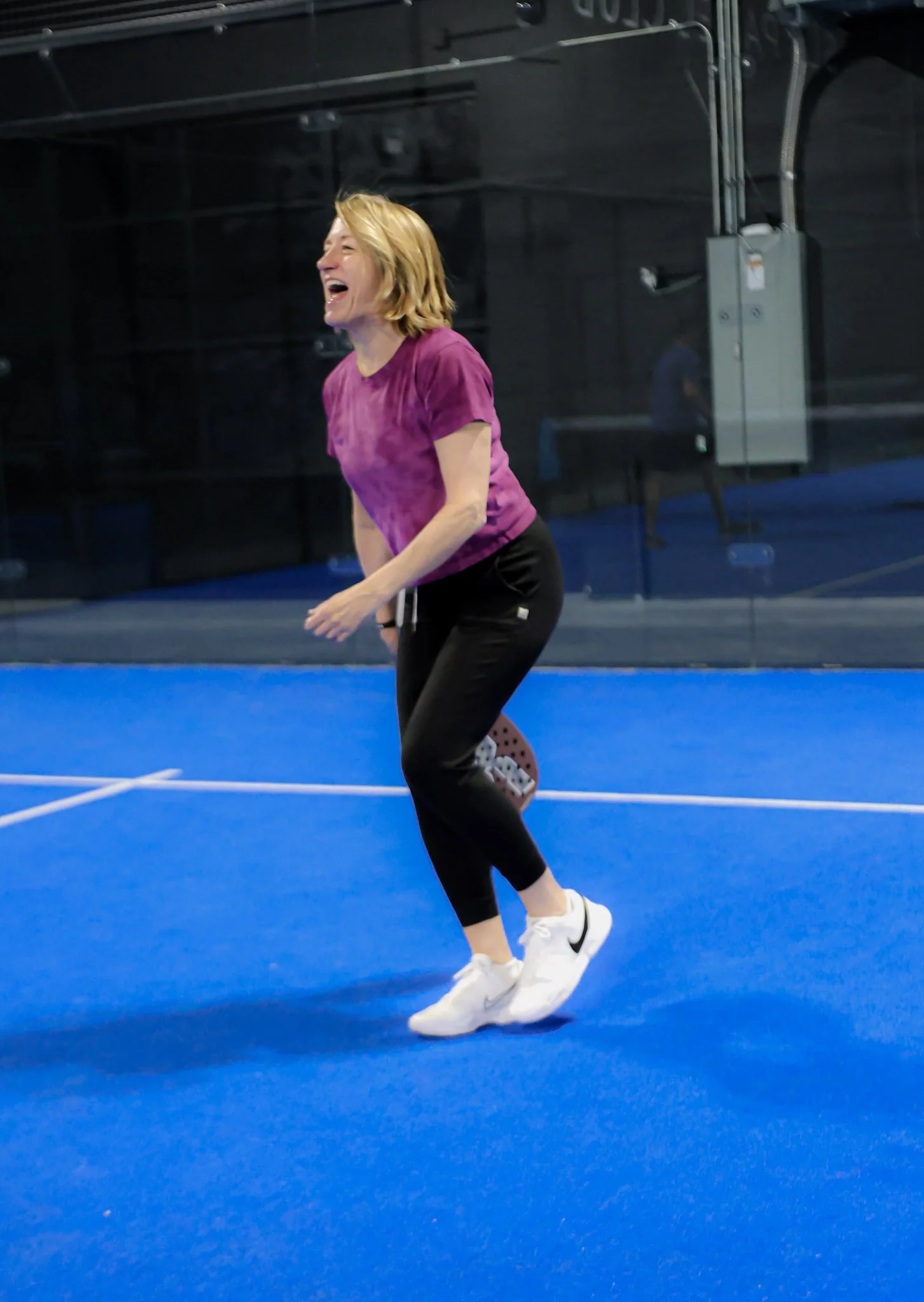A woman laughing and playing padel in a profitable padel club, wearing a purple shirt, black pants, and white sneakers.