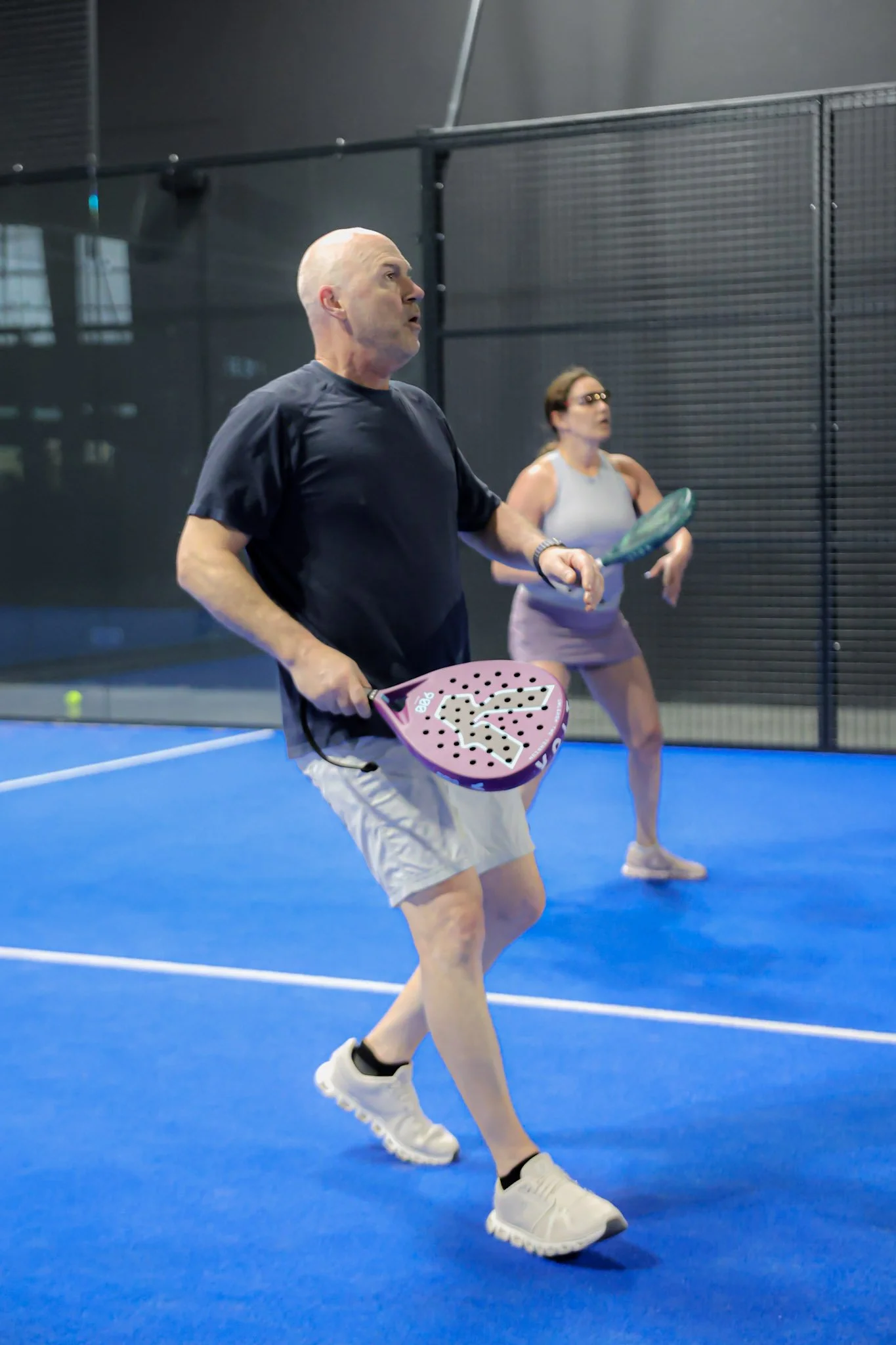 A man and a woman playing padel indoors on a blue padel court, holding padel racquets, with a black net behind them.