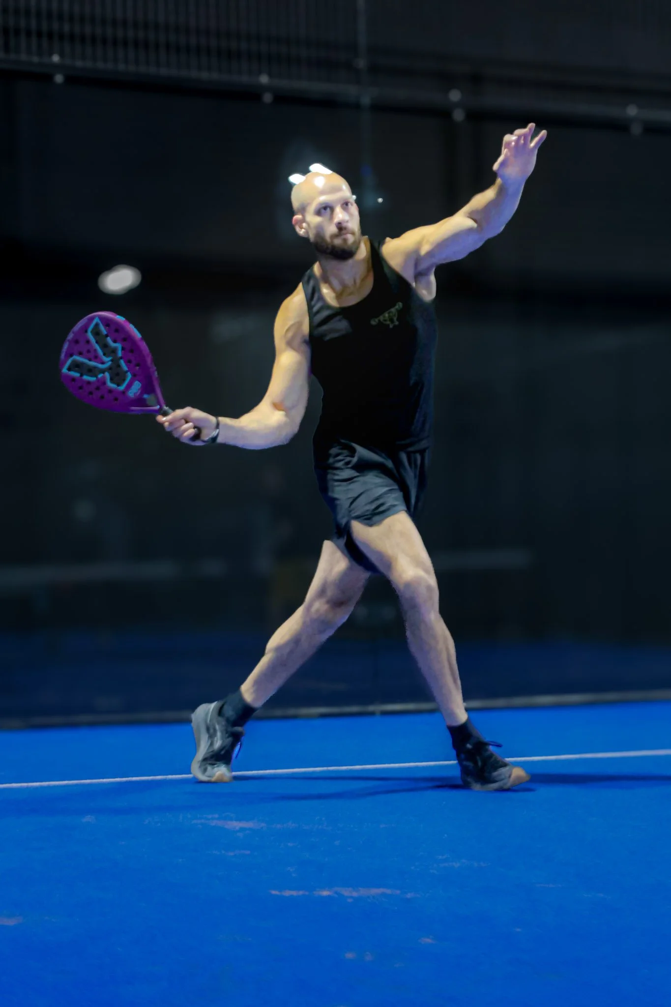 A man playing padel on an indoor padel court, holding a padel racquet, wearing black athletic gear, in a dynamic stance.