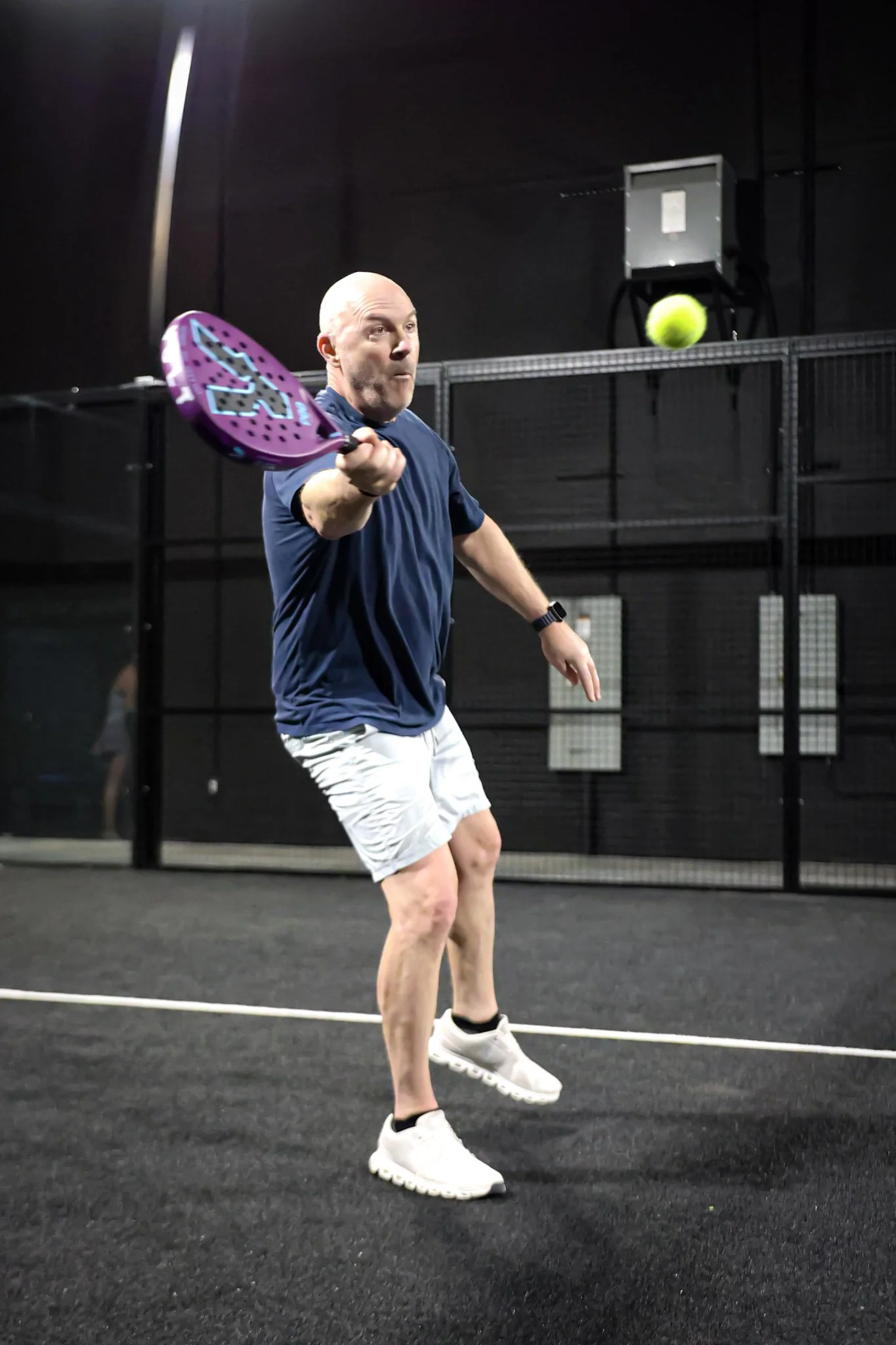 Man playing padel on indoor padel court, about to hit the ball with athletic stance, wearing navy shirt, white shorts, and white sneakers.