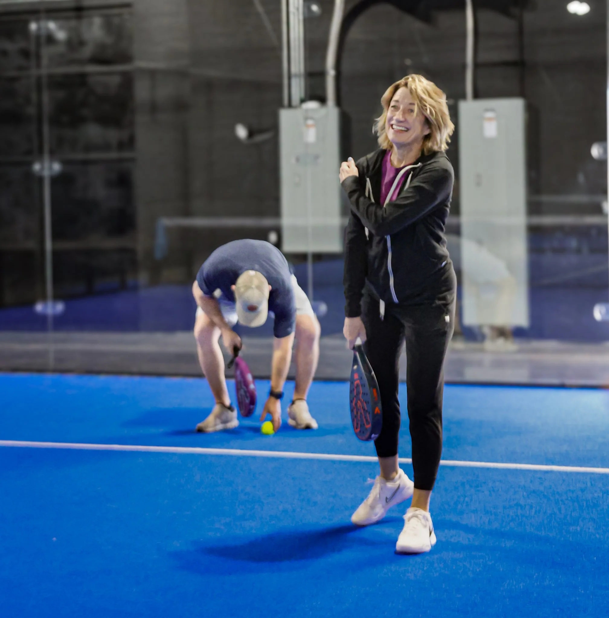 A woman with blond, wavy hair smiling and standing on a blue padel court. There is padel equipment provided by Peak Padel in the background
