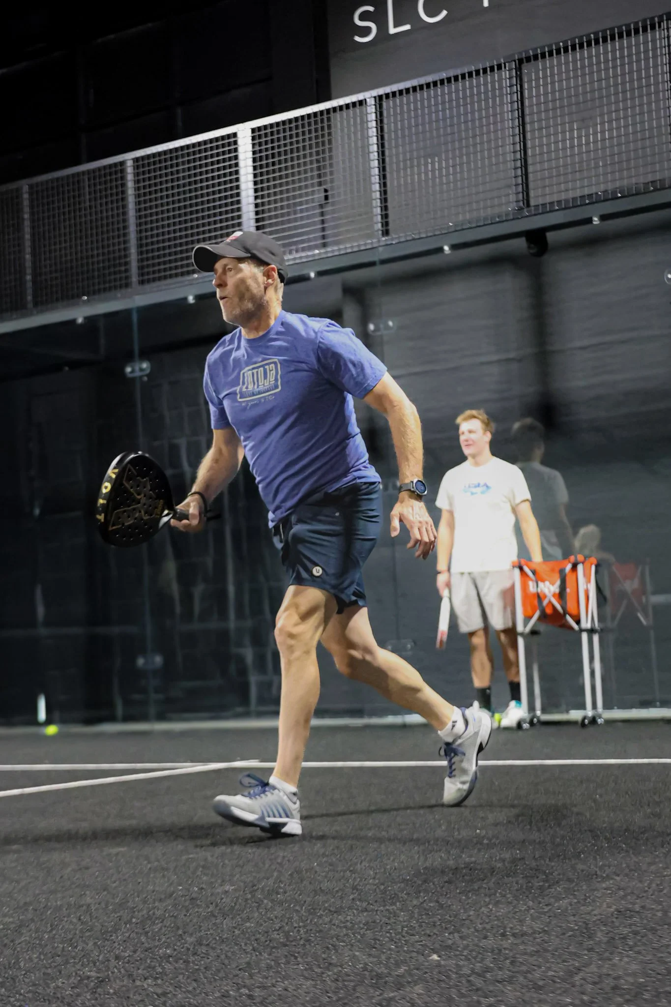 A man jogging on an indoor padel court holding a padel racquet, with several padel balls and a cart of padel balls nearby in the background. Another person is standing further back on the padel court.