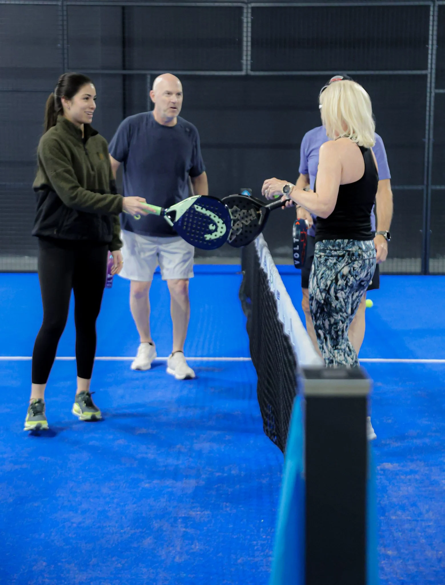 Four people standing on a blue padel court, engaged in conversation. One woman is dressed in a black athletic dress with patterned leggings and blonde hair, and the other woman in a green jacket with black leggings and dark hair. 