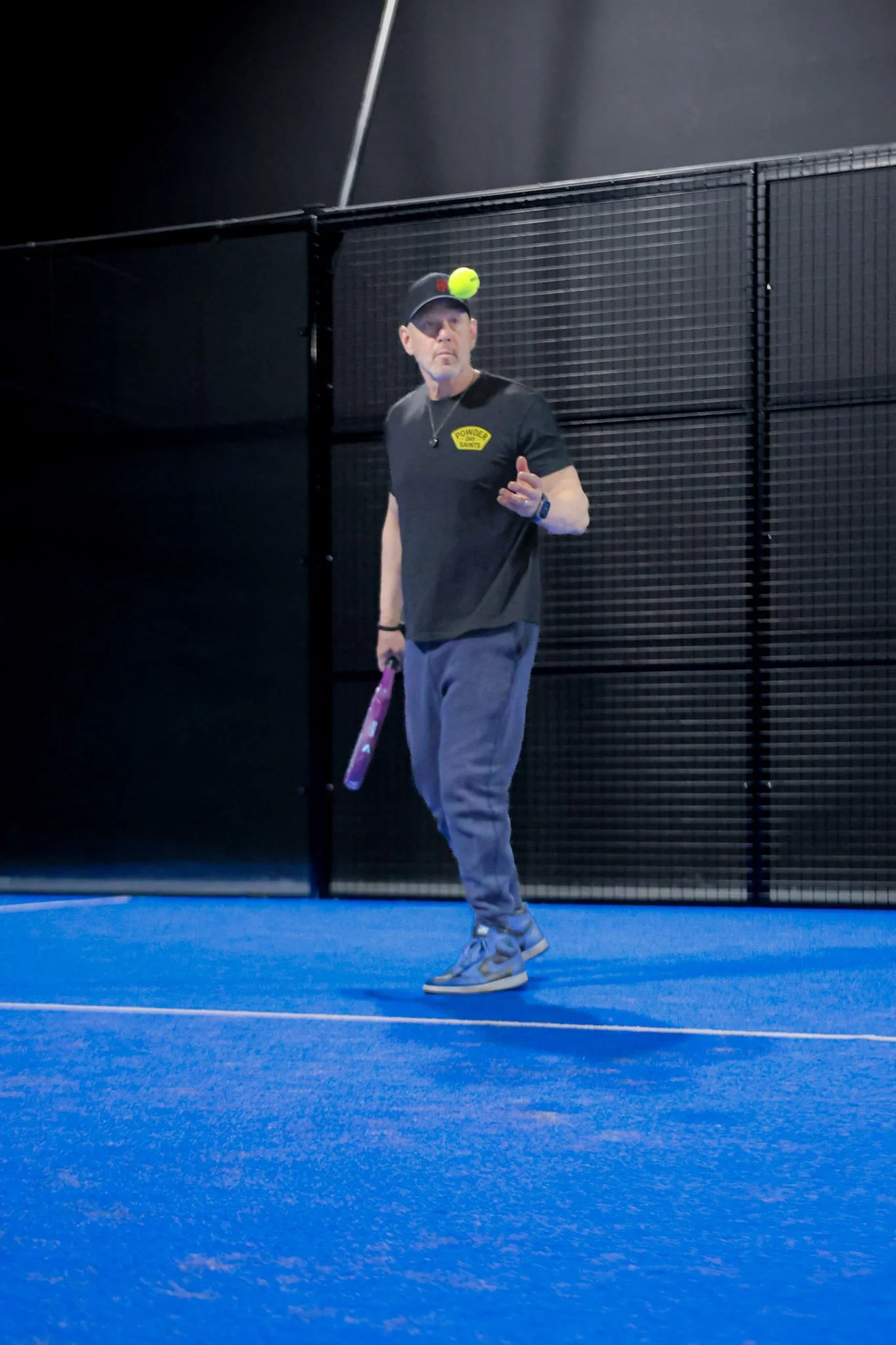 A man standing on a blue indoor padel court, ready to hit a padel ball, with a black fence in the background.