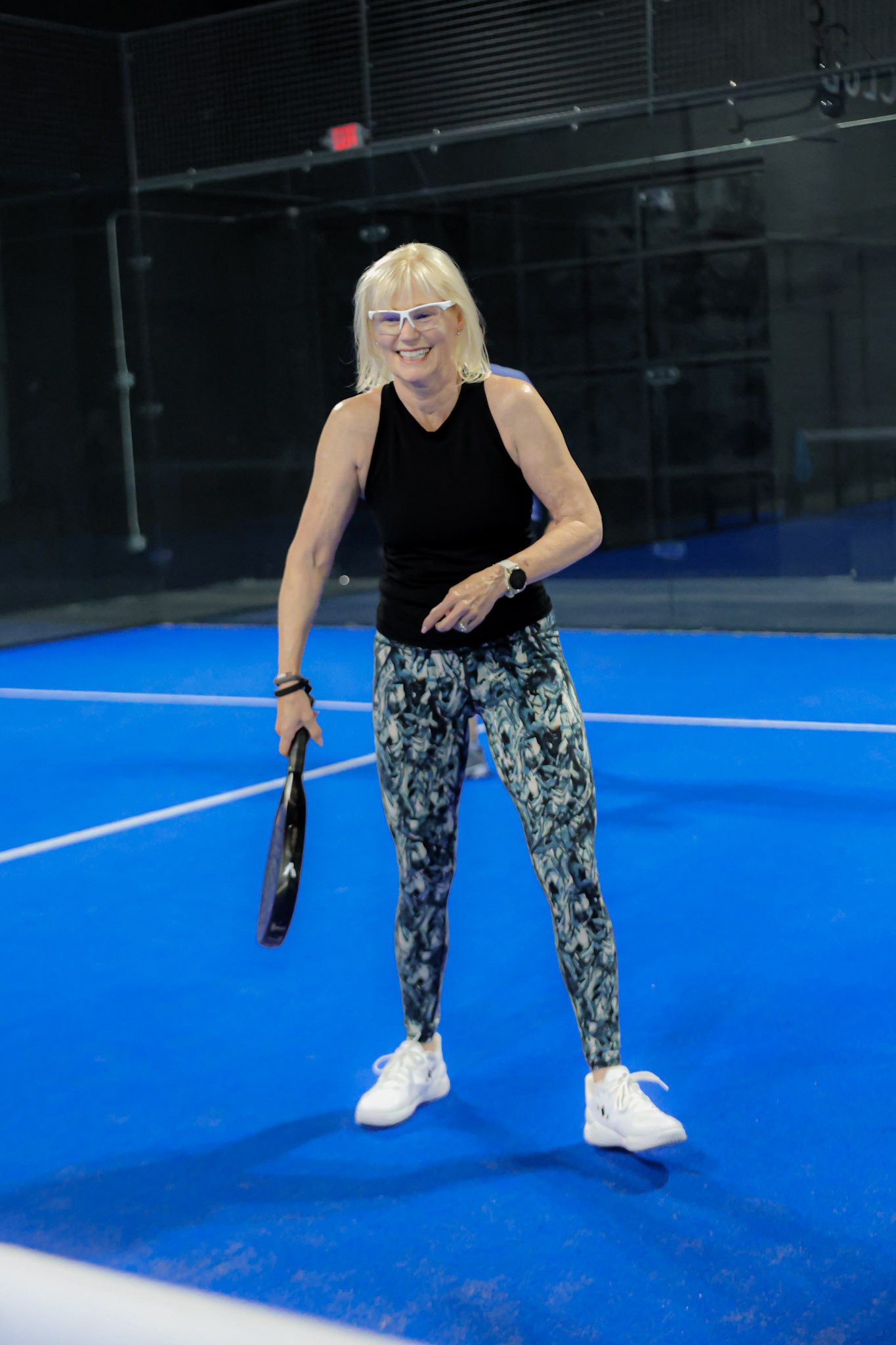 A smiling woman in athletic clothing holding a padel racket on an blue padel court.