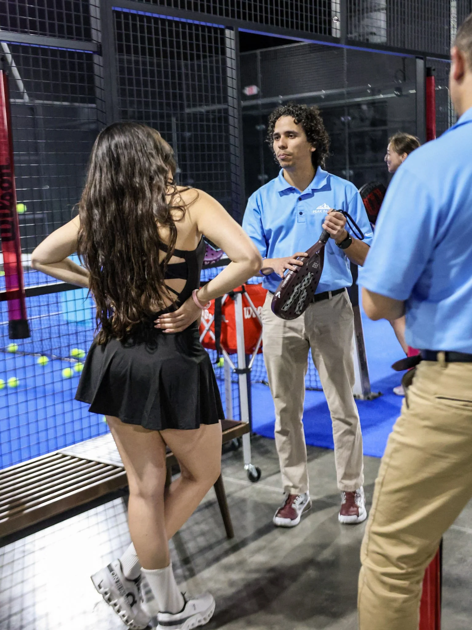 A young woman with long, wavy hair in a black dress and white sneakers is talking to a man in a blue polo and beige pants in a successful padel club. The woman is leaning against a fence with padel balls behind her. 