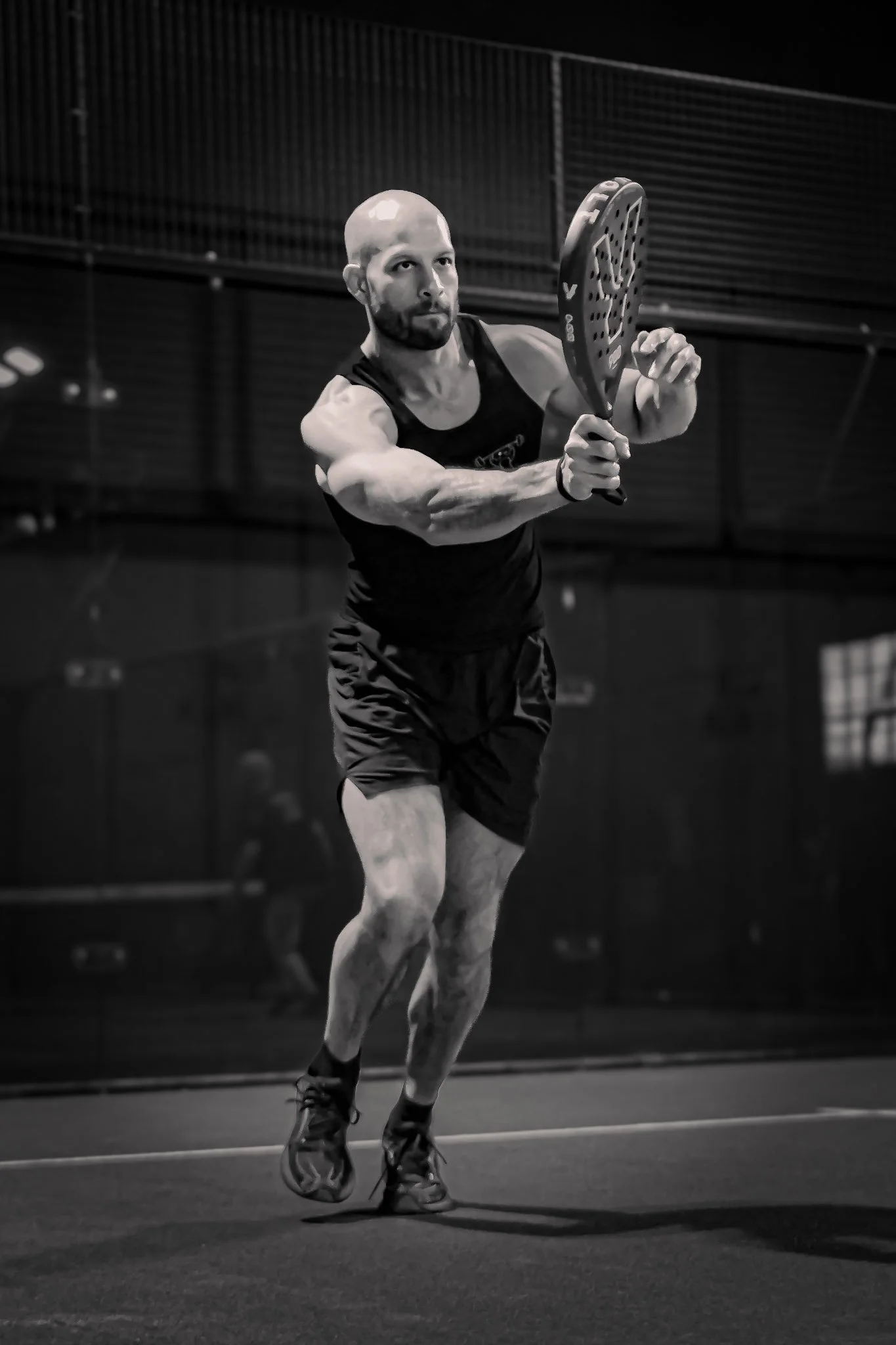 A black and white photo of a man playing padel tennis, preparing to hit the ball with his padel racket on an outdoor padel court at night.