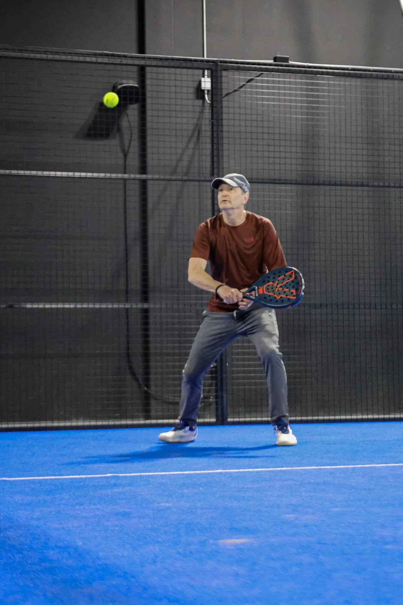 An older man playing padel on an indoor padel court with a blue surface, wearing a gray cap, red t-shirt, gray pants, and white shoes, holding padel racquet and preparing to hit a neon yellow padel ball.