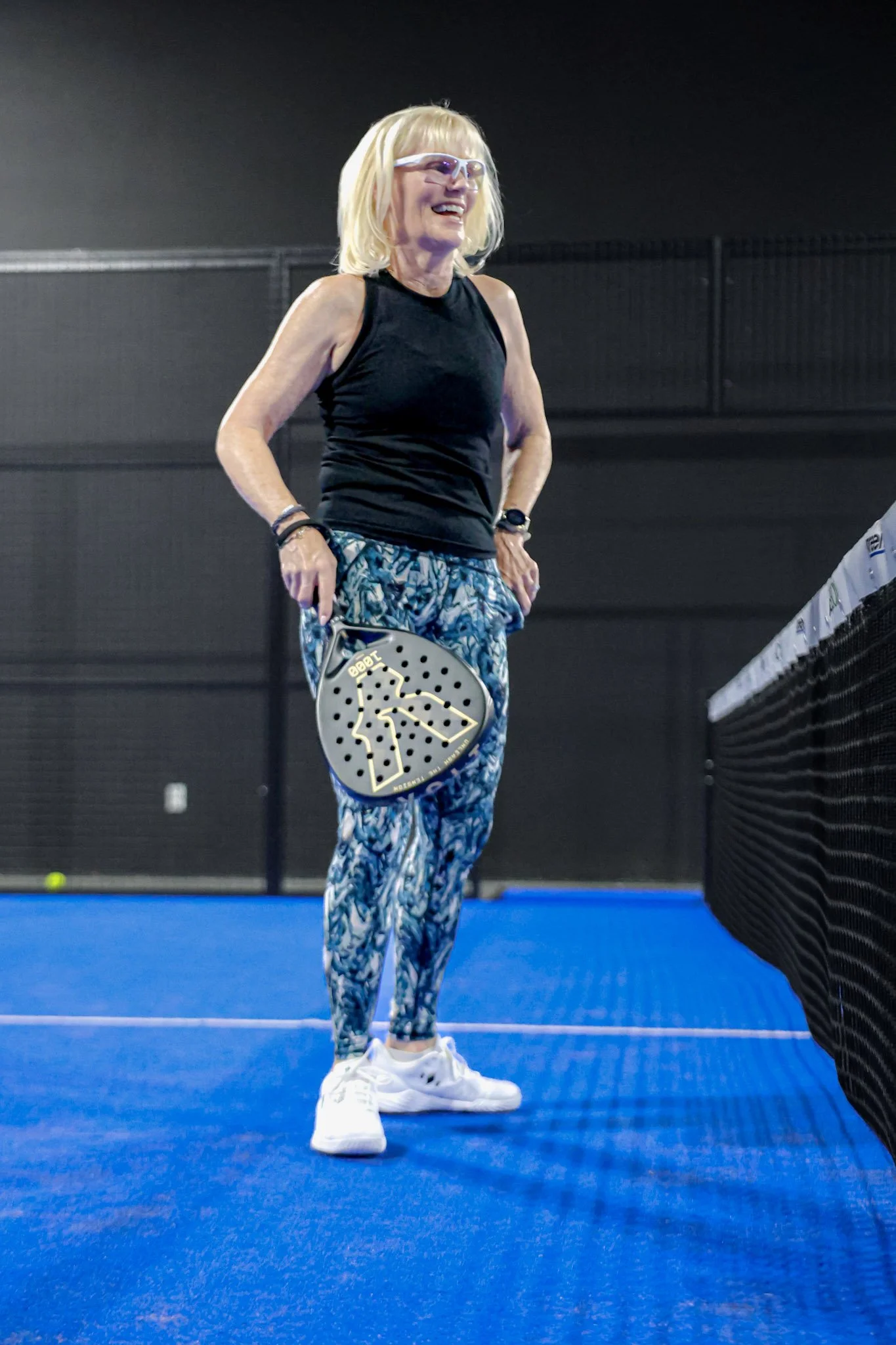 Middle-aged woman with blonde hair, wearing glasses, black sleeveless top, patterned blue athletic pants, and white sneakers, standing on a blue padel court holding a padel racquet during a game.