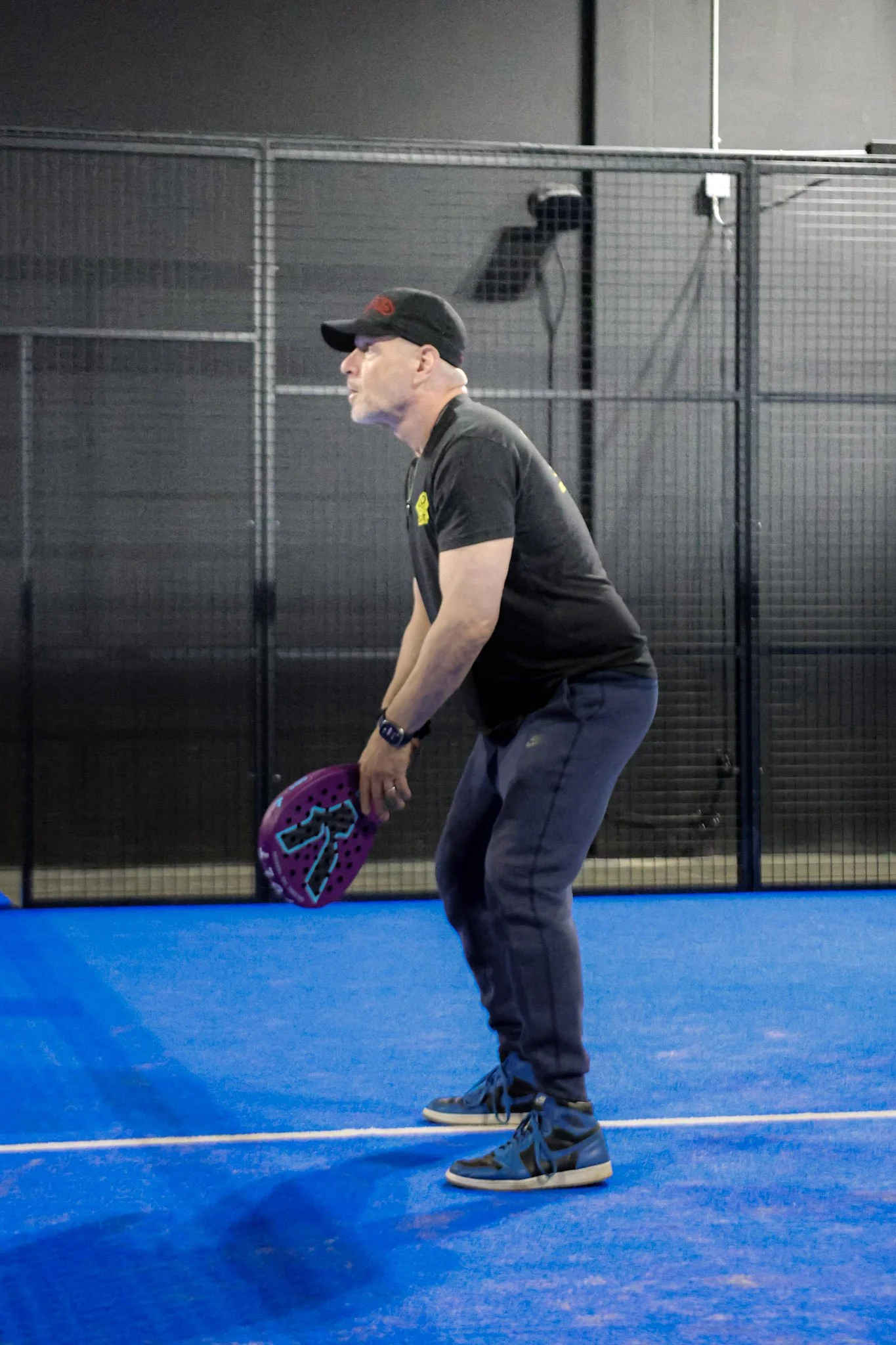 A man in gray pants, black shirt, and a black cap holding a purple padel racquet stands on a blue indoor padel court, with a black fence and a person in the background.