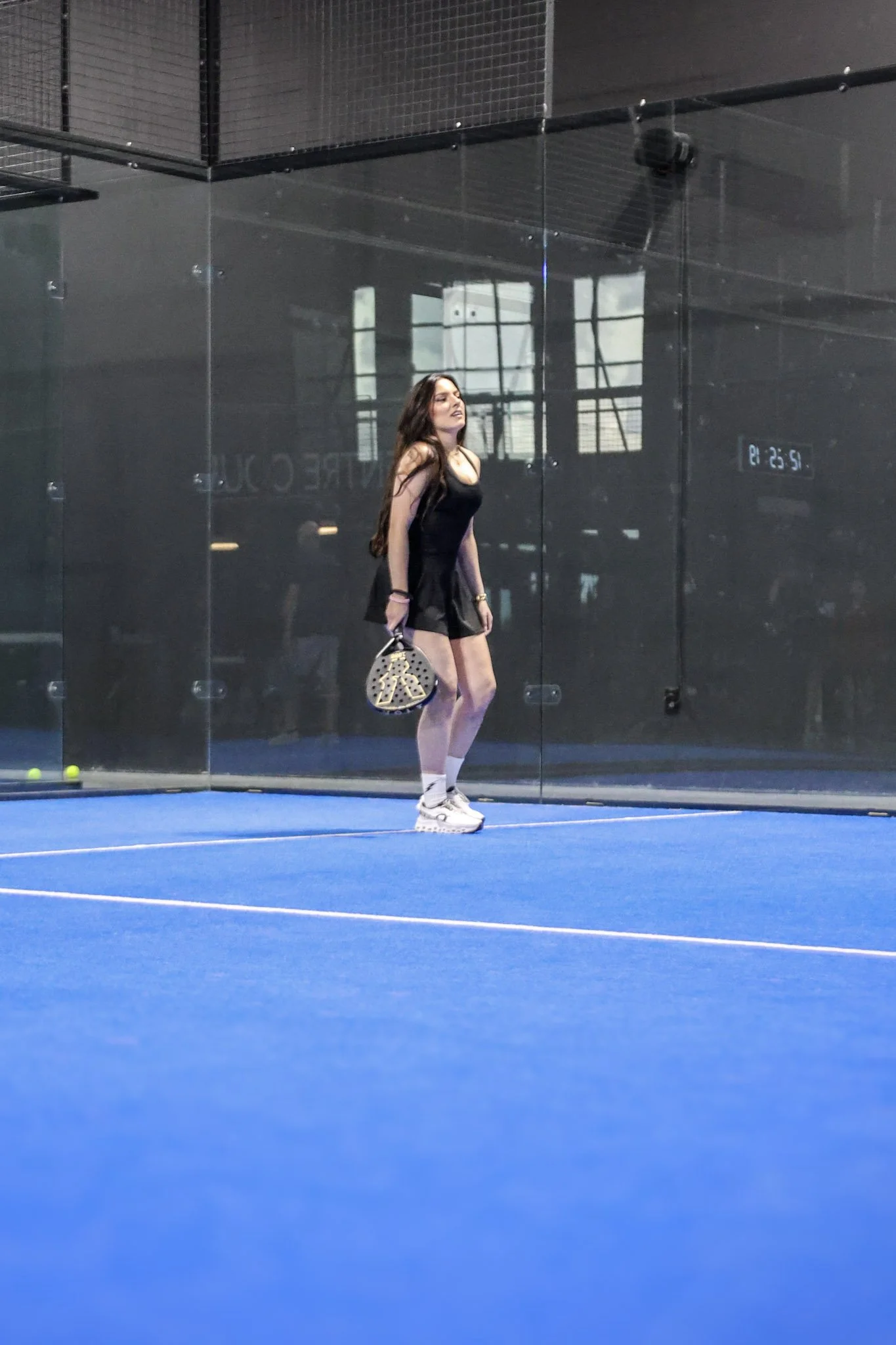 A woman with long dark hair in a black dress and white sneakers standing on a blue indoor padel court, holding a padel racquet and looking to her left. There is a glass wall behind her with reflections and a digital timer on the wall.