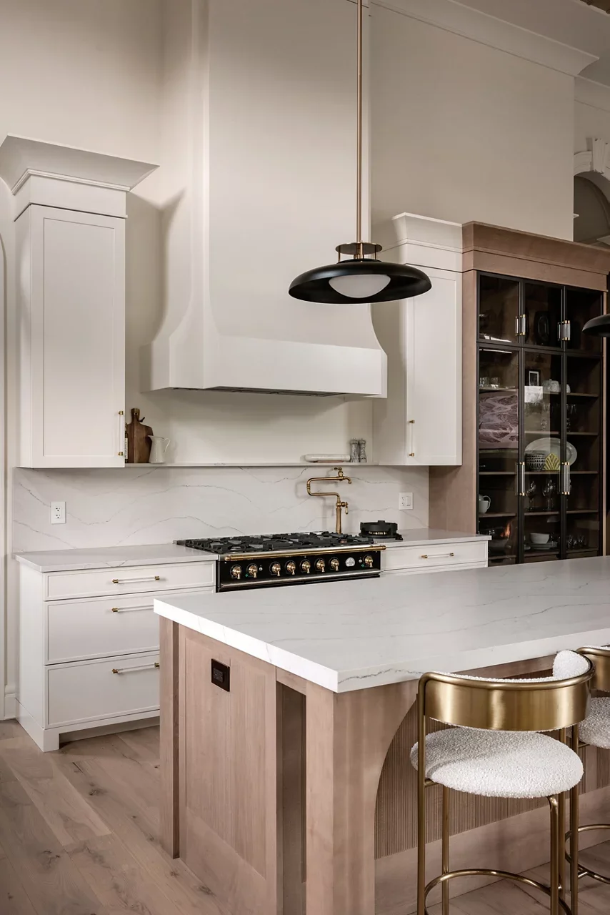 Modern kitchen with white cabinets, a marble island, a black and gold stove, a hanging black pendant light, and a glass-front wooden cabinet filled with dishes.