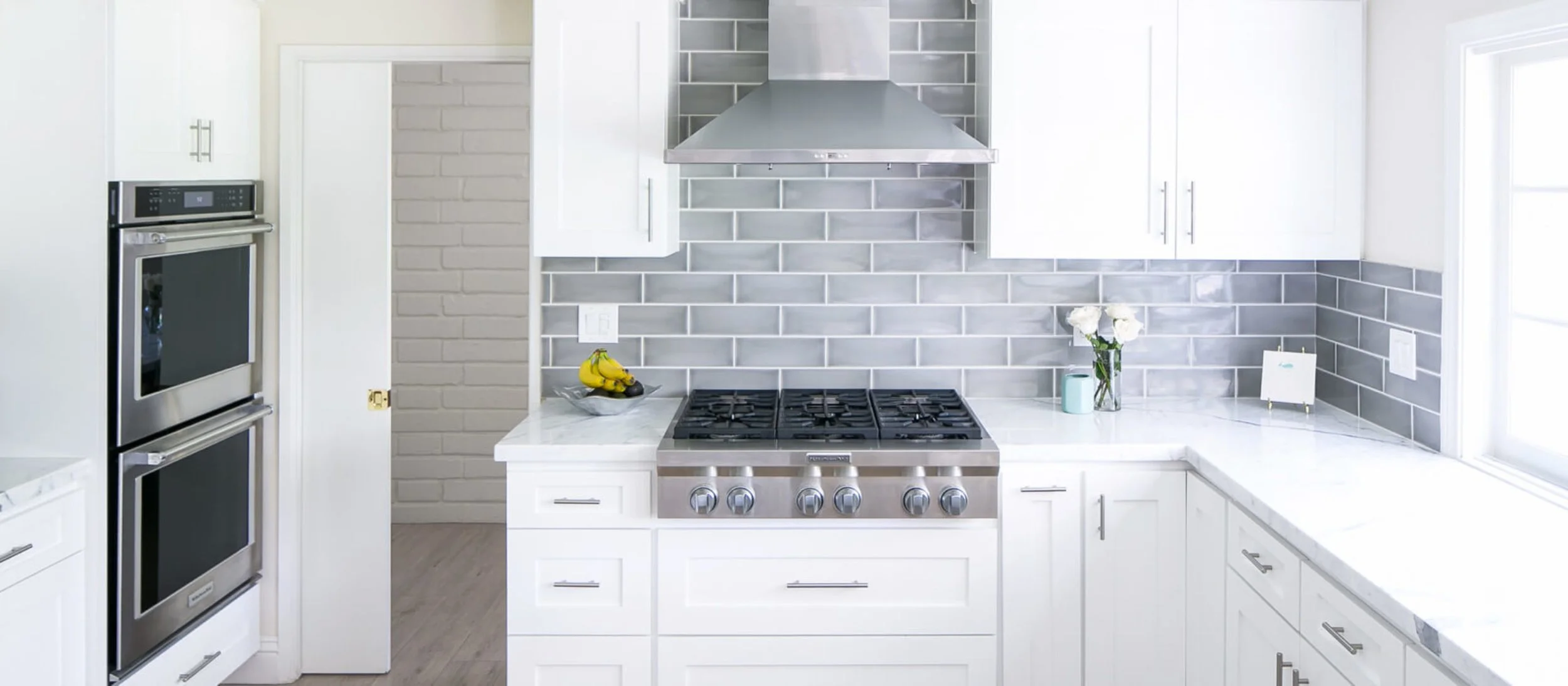 Modern kitchen with white cabinets, gray subway tile backsplash, stainless steel range and oven, yellow bananas on a black plate, flowers in a vase, and light streaming through windows.