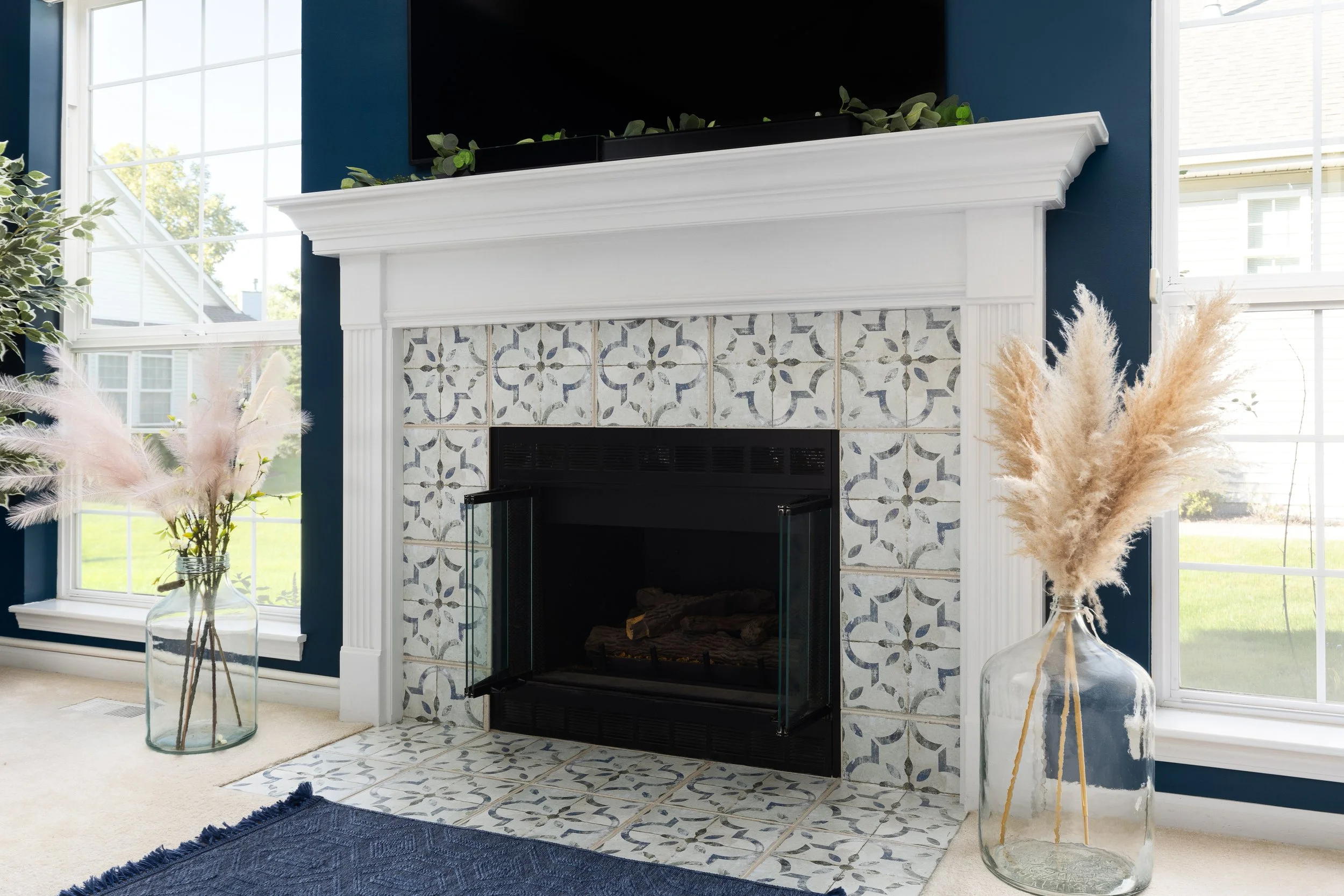 Living room with a white painted fireplace surrounded by patterned tiles, large windows on both sides, and decorative vases with pampas grass on either side of the fireplace.