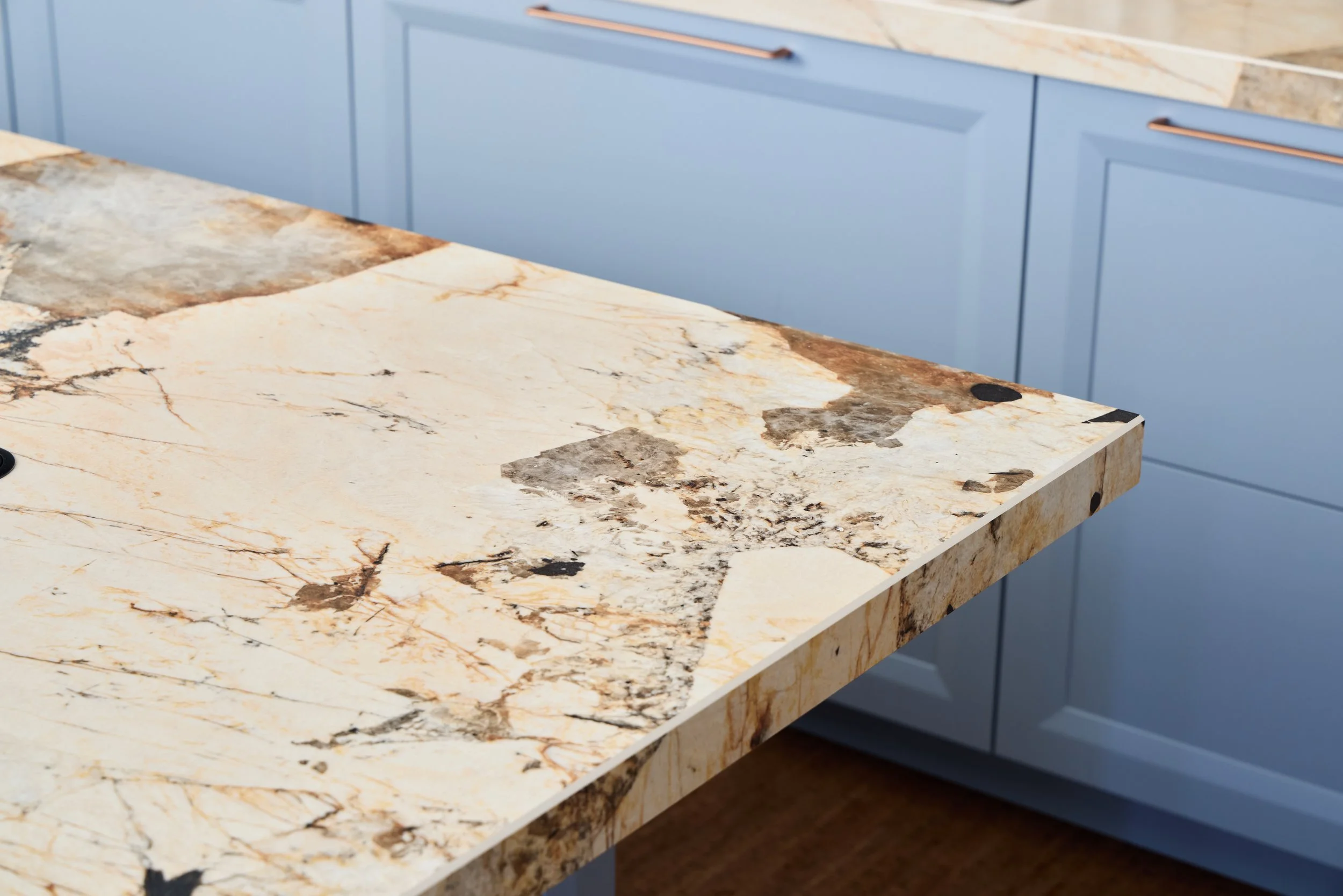 Close-up of a polished granite kitchen countertop with cream, brown, and black veining, adjacent to blue cabinet doors with gold handles.