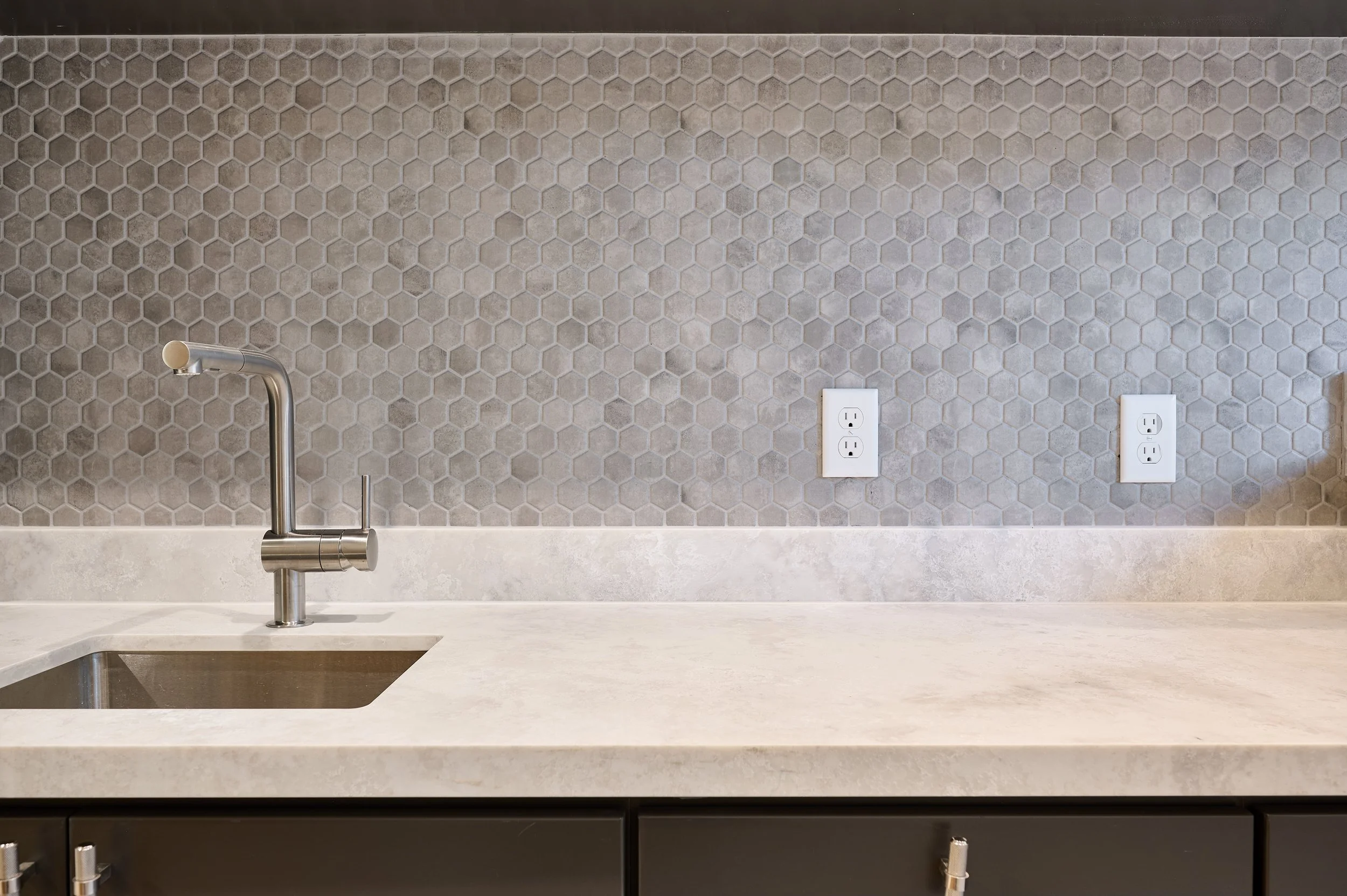 Modern kitchen countertop with stainless steel faucet, light gray hexagonal tile backsplash, two white electrical outlets, and black cabinet drawers.