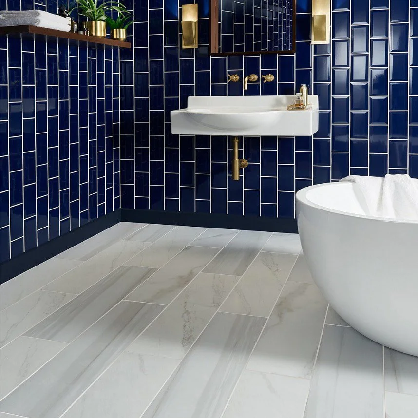 Modern bathroom with navy blue tiled walls, a white wall-mounted sink with gold fixtures, a white oval-shaped bathtub with a towel inside, and beige floor tiles with gray veining.