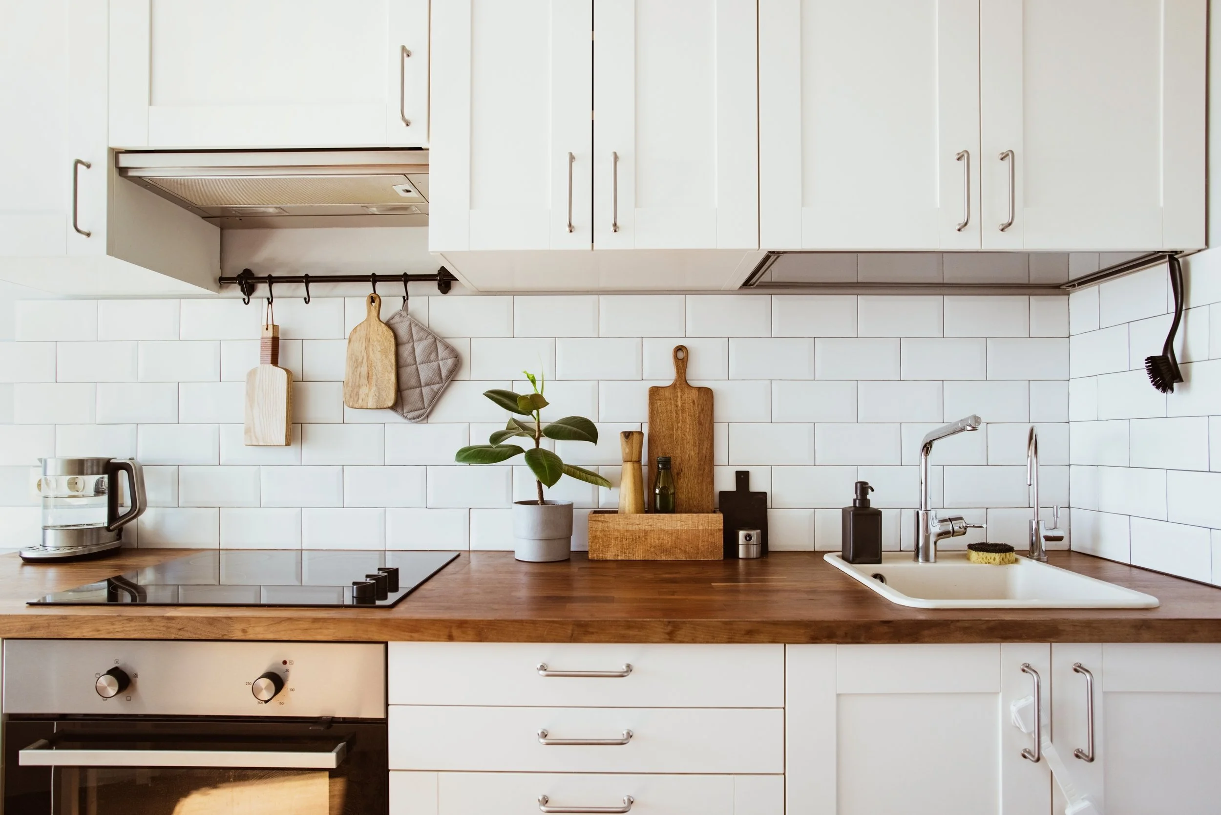Modern kitchen with white cabinets, wooden countertop, and white subway tile backsplash. Contains a stove, a coffee maker, a potted plant, cutting boards, and kitchen utensils.