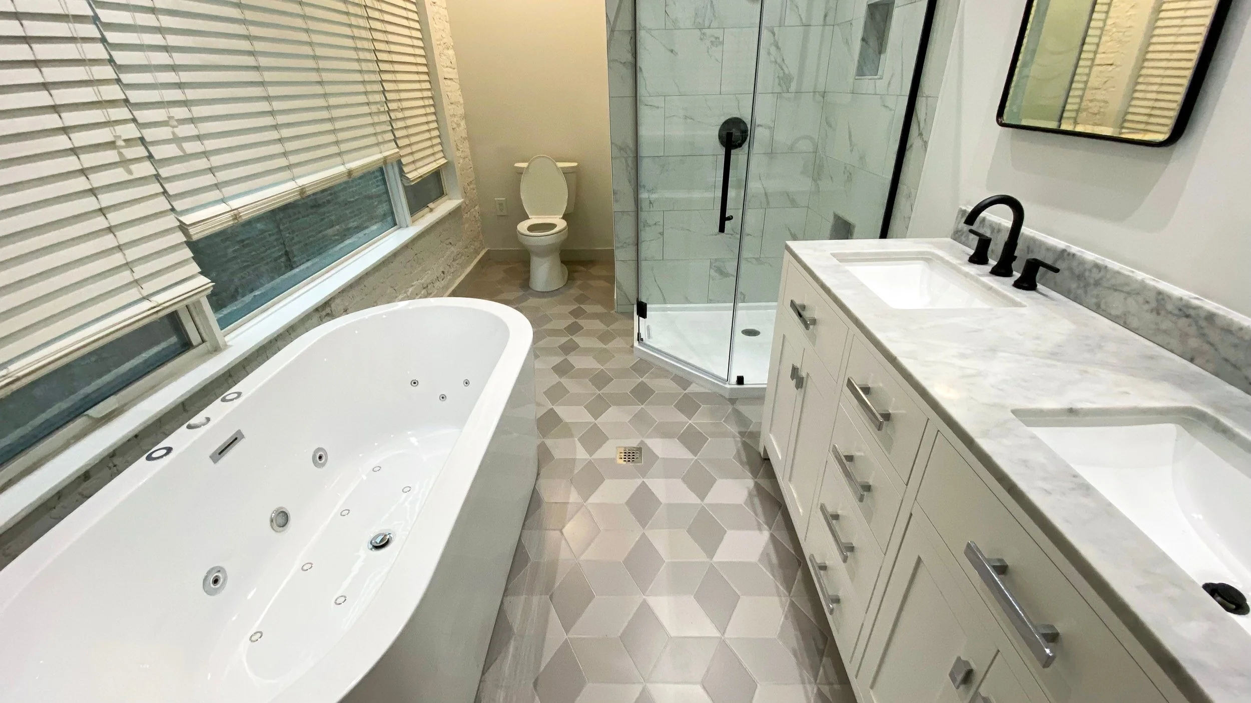 Modern bathroom with a large bathtub, a glass-enclosed shower, a toilet, and a white vanity with dual sinks and black fixtures, accented by hexagonal floor tiles.