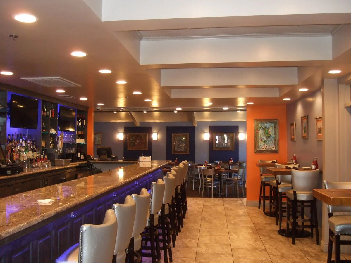 Inside a modern bar and restaurant with a long marble counter, cream-colored bar stools, and small tables with chairs. Canda Construction commercial construction and renovation.