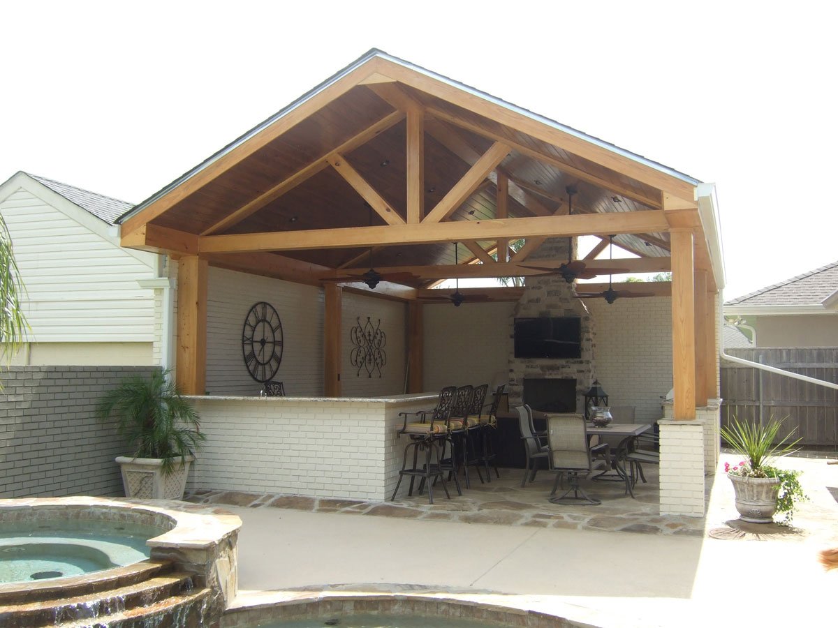 Outdoor patio with wooden roof structure, ceiling fans, and a fireplace, surrounded by a white brick wall, with outdoor furniture and potted plants, in a backyard setting. Canda Construction residential construction and renovation.