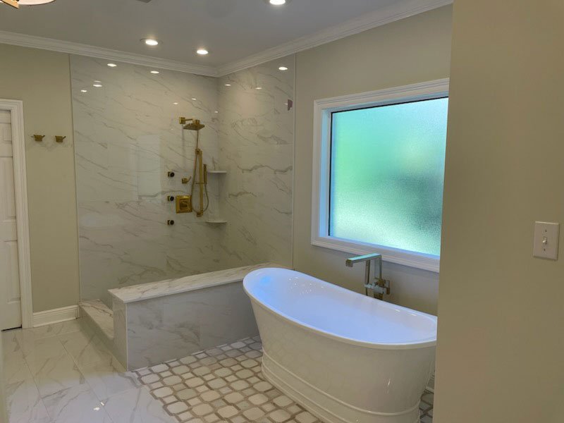 A modern bathroom with a freestanding oval bathtub near a window with frosted glass, and a walk-in shower area with marble walls, a gold showerhead, and built-in shelves. Canda Construction residential construction and renovation.
