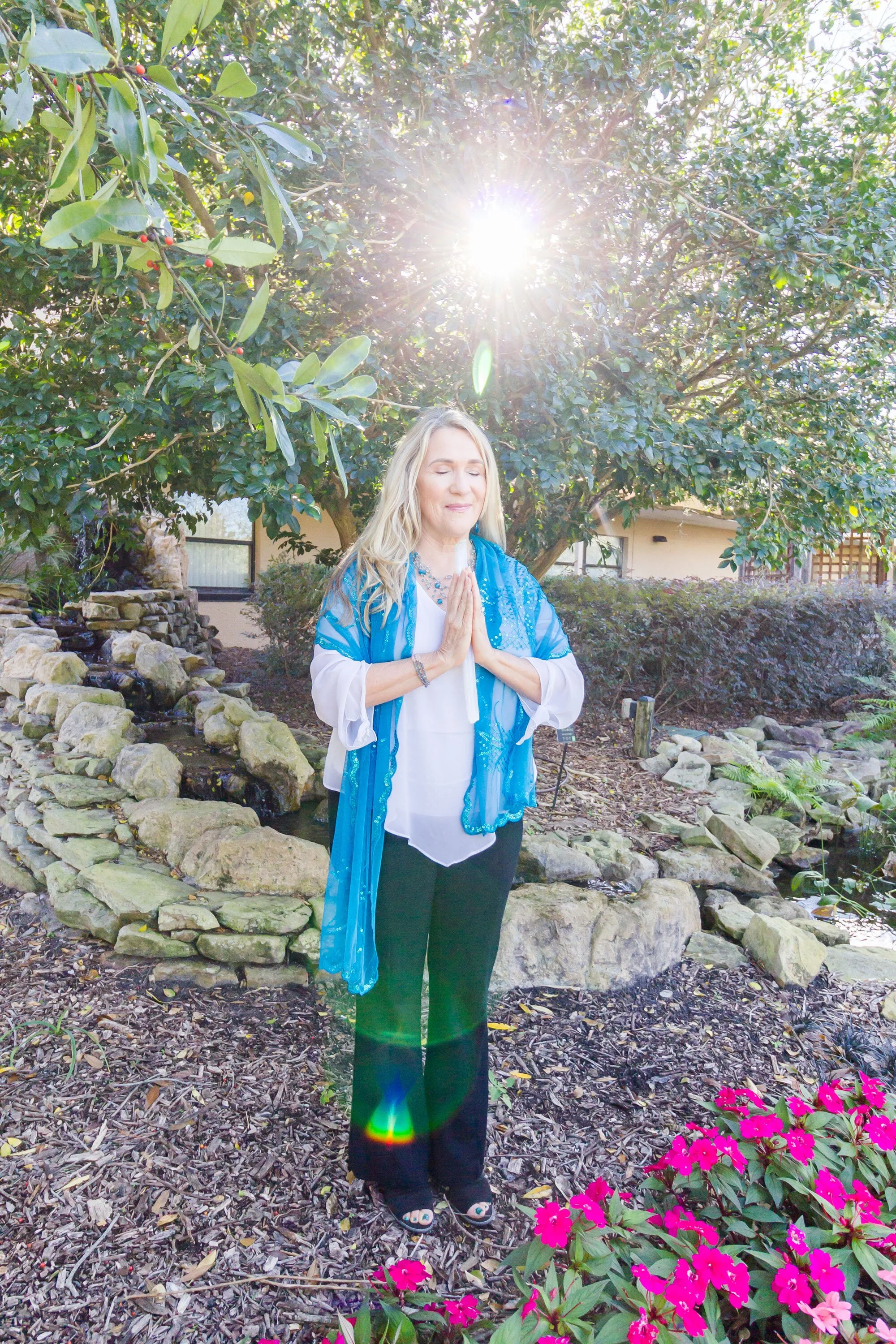 Rev. Dr. Debbi Adams standing beneath a tree with hands at heart center and eyes closed as sunlight shines above her