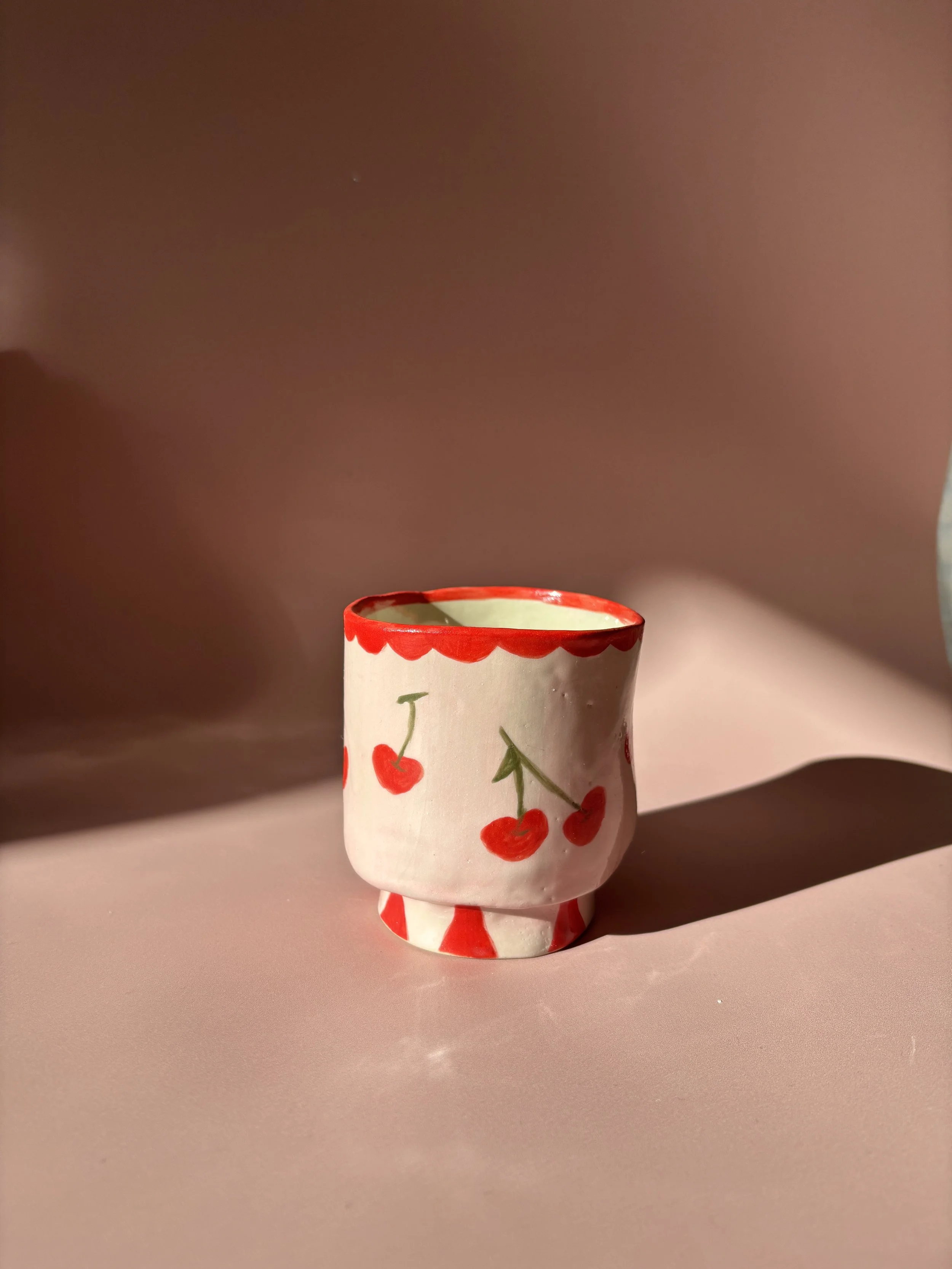 A small ceramic cup with a cherry design, featuring red cherries and green stems, on a pink surface with a soft pink background.