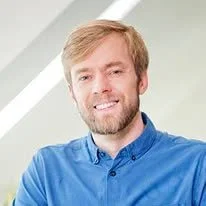 A man with light brown hair and a beard, wearing a blue collared shirt, smiling in front of a light-colored background.