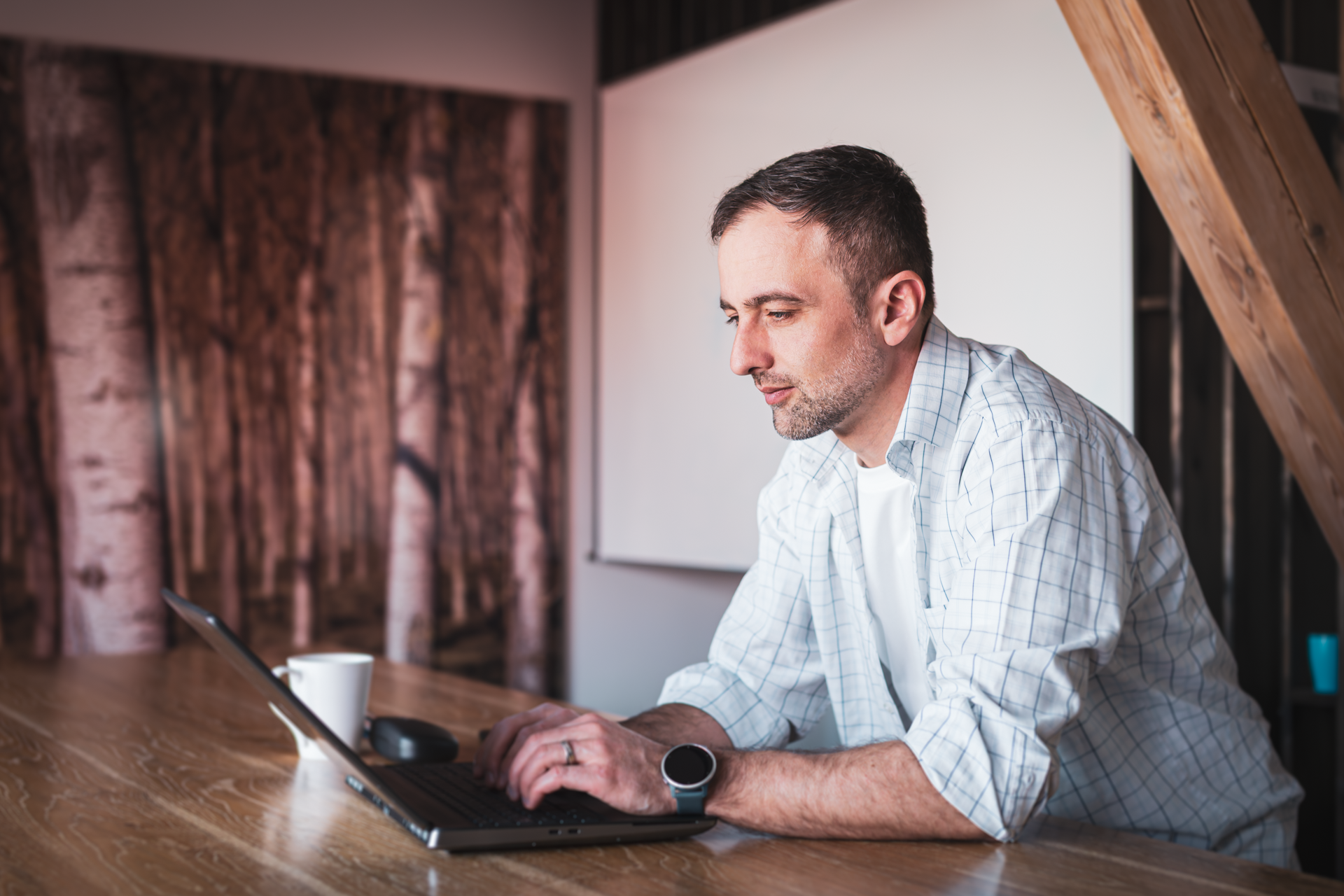 Tibor Kadar, wearing a plaid shirt, working on a laptop at a wooden table in a modern office space.