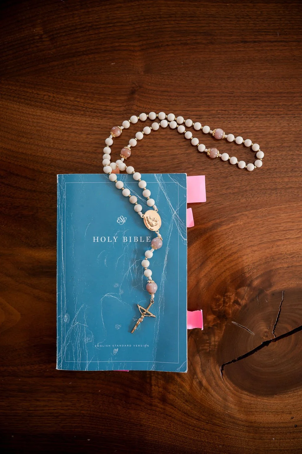 A rosary with white and pink beads, a crucifix, and an image of a saint, resting on a blue Holy Bible with pink ribbons on a wooden surface.