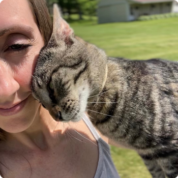 A woman with a gray striped tabby cat hugging her cheek outdoors