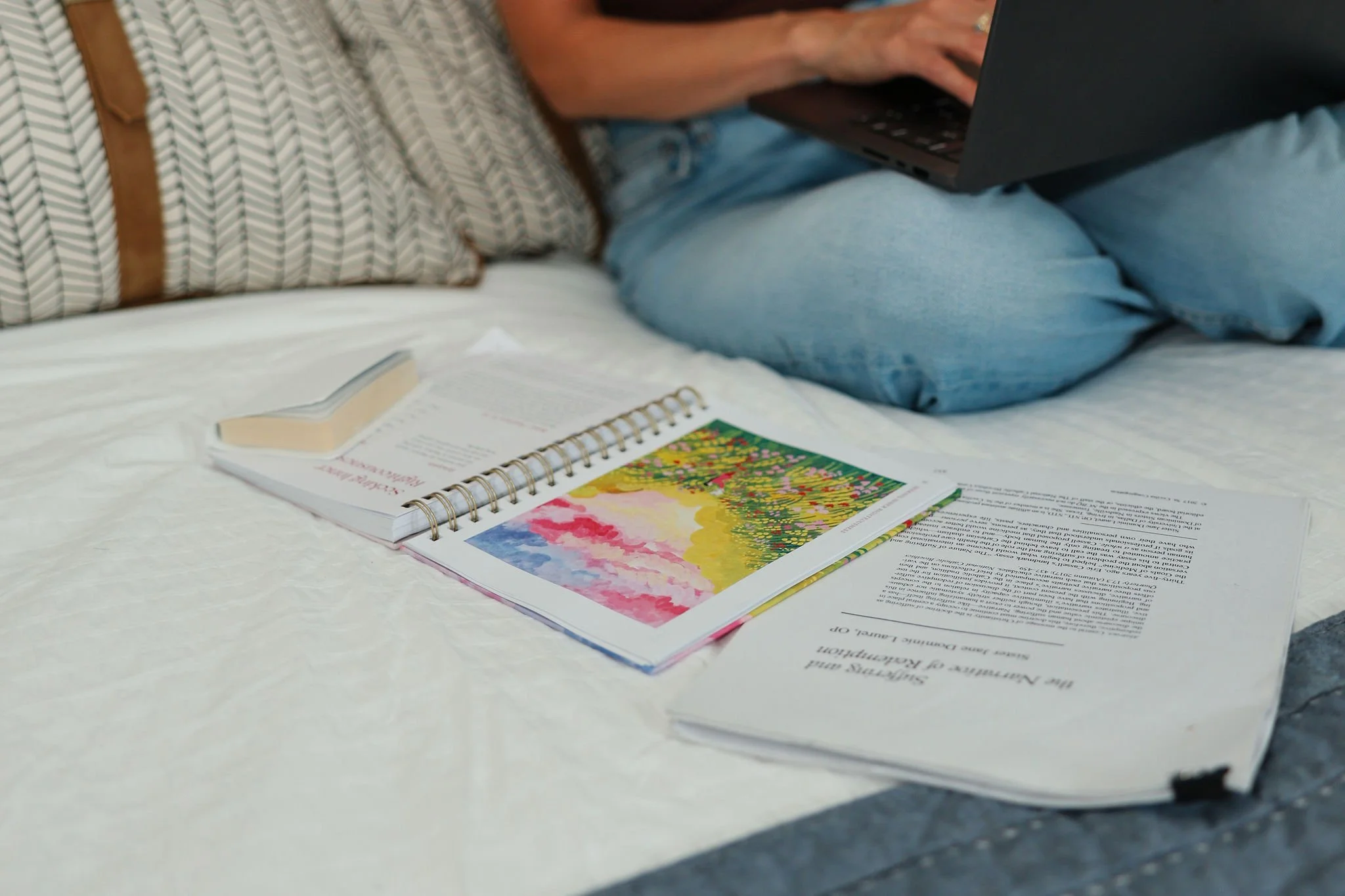 Person sitting on a bed using a laptop, with open notebook, printed documents, and a colorful illustrated book spread out on the bed.