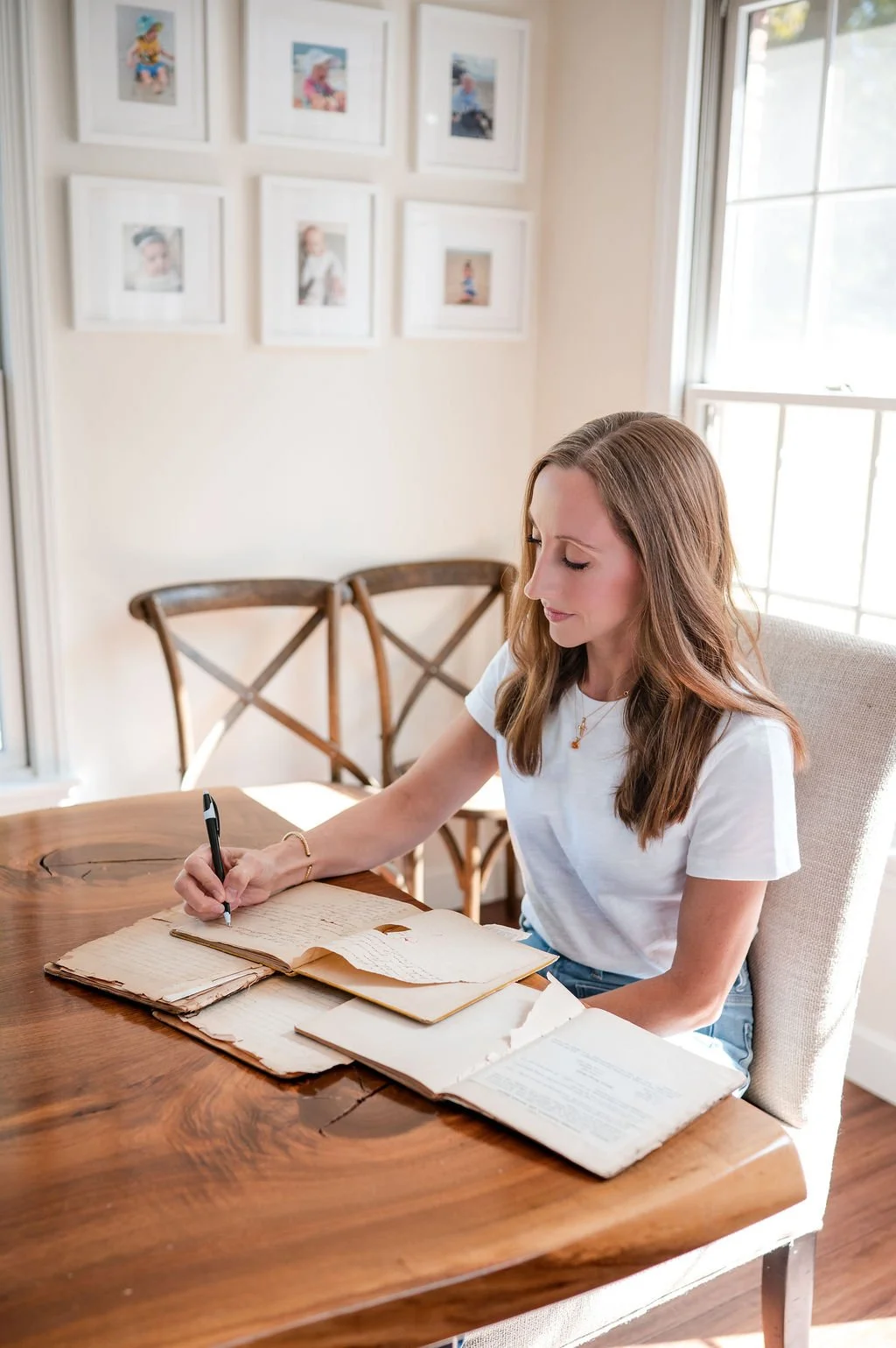 A woman with brown hair and a white t-shirt sitting at a wooden table, writing in an old, open book with other aged papers on the table. The room has cream walls, a window letting in natural light, and framed photos on the wall.