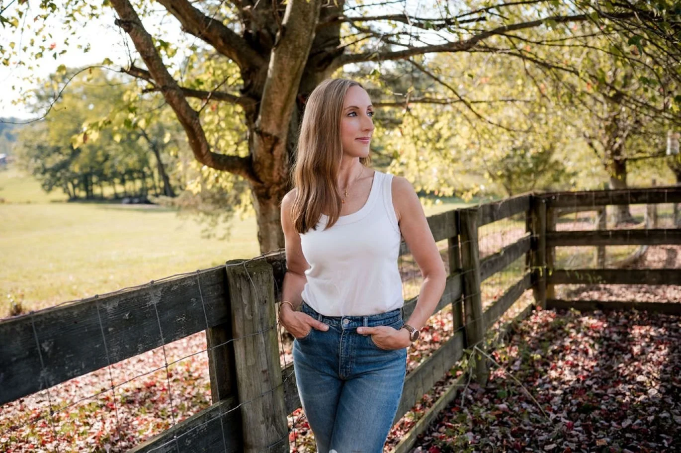 A woman in white top and jeans standing by a wooden fence in a park during fall, with trees and fallen leaves in the background.