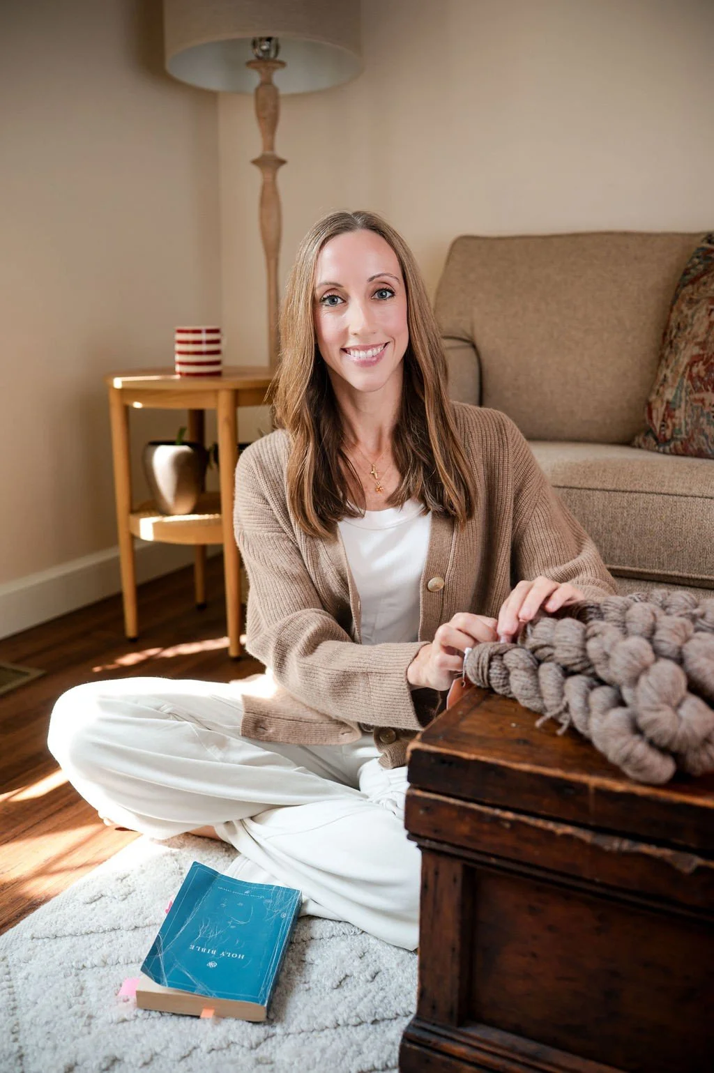 A woman sitting on the floor in a cozy living room, looking at the camera and smiling, with a book on the rug and a chunky knit blanket on a wooden chest.