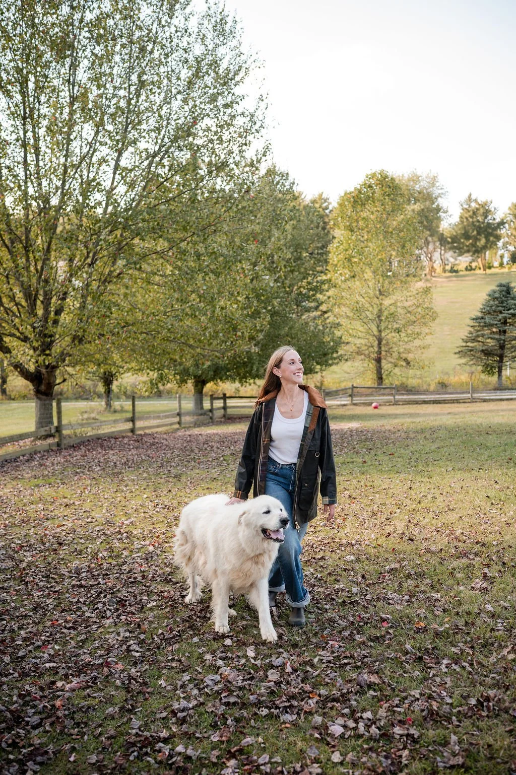 A woman walking outdoors in a park with a large, fluffy white dog, surrounded by trees with green leaves and a grassy landscape.