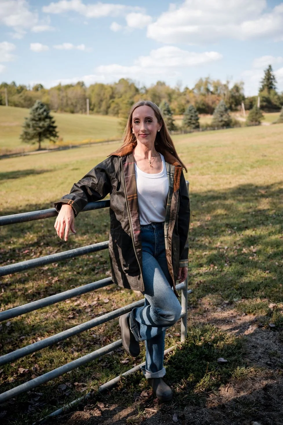 A woman standing outdoors by a metal fence in a grassy field with trees in the background and a partly cloudy sky.